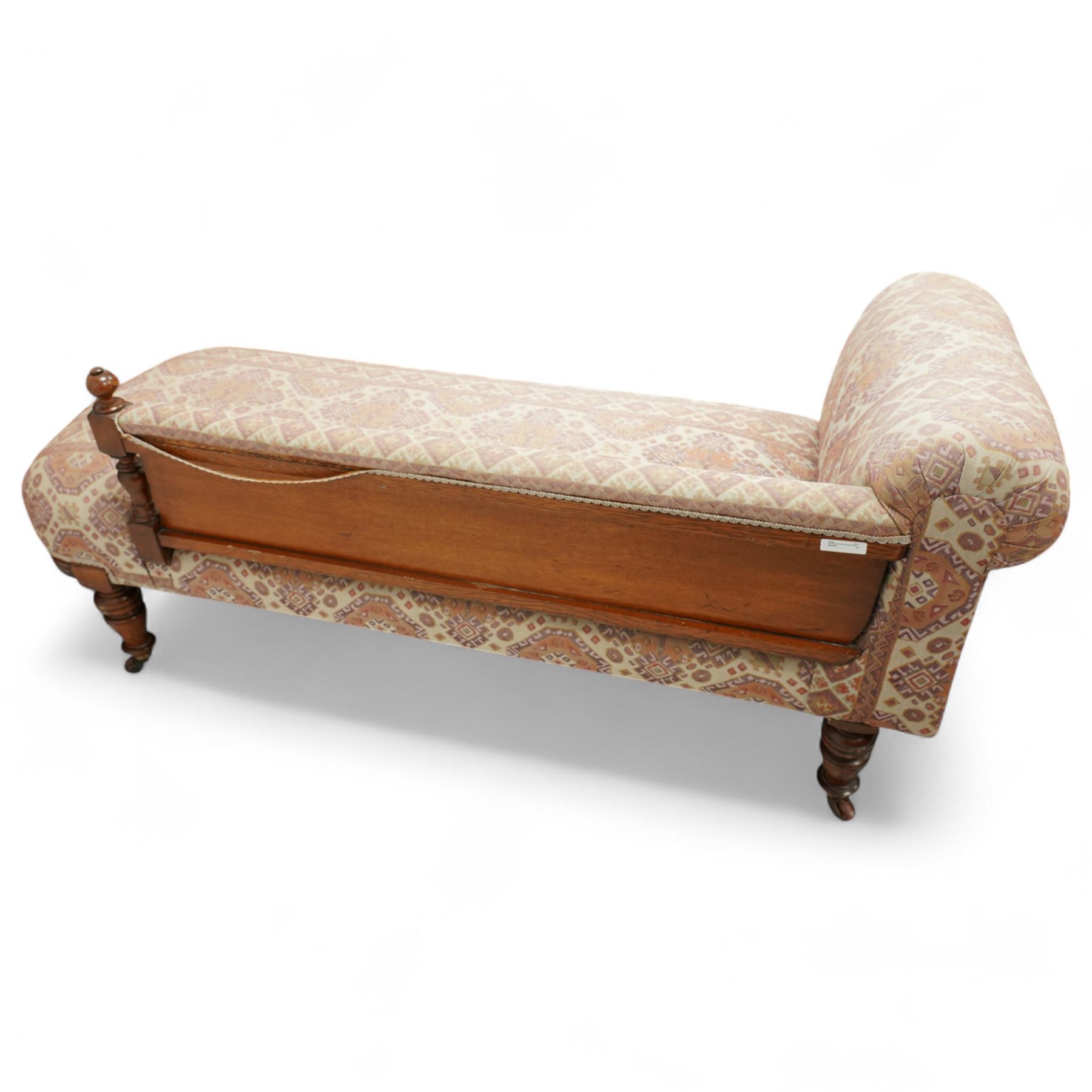 Late Victorian walnut chaise longue, upholstered in geometric patterned fabric, the scroll end and padded back above carved side panel with foliate and roundel motifs, shaped seat rail on turned front supports with ceramic castors