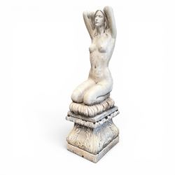 20th century composite stone garden figure, modelled as a kneeling nude female with arms raised behind her head, mounted on an integral cushion, the squared ogee pedestal moulded with acanthus leaves and flower heads, in a white painted and weathered finish