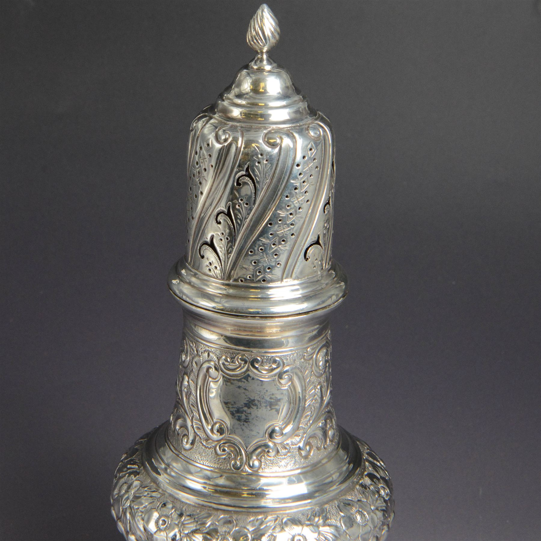 Victorian silver sugar caster, of bellied urn form, embossed with floral and foliate motifs, with two vacant cartouche, upon a domed stepped circular foot, with pierced removable cover with flambe finial, hallmarked Marston & Bayliss, Chester 1900, H25cm