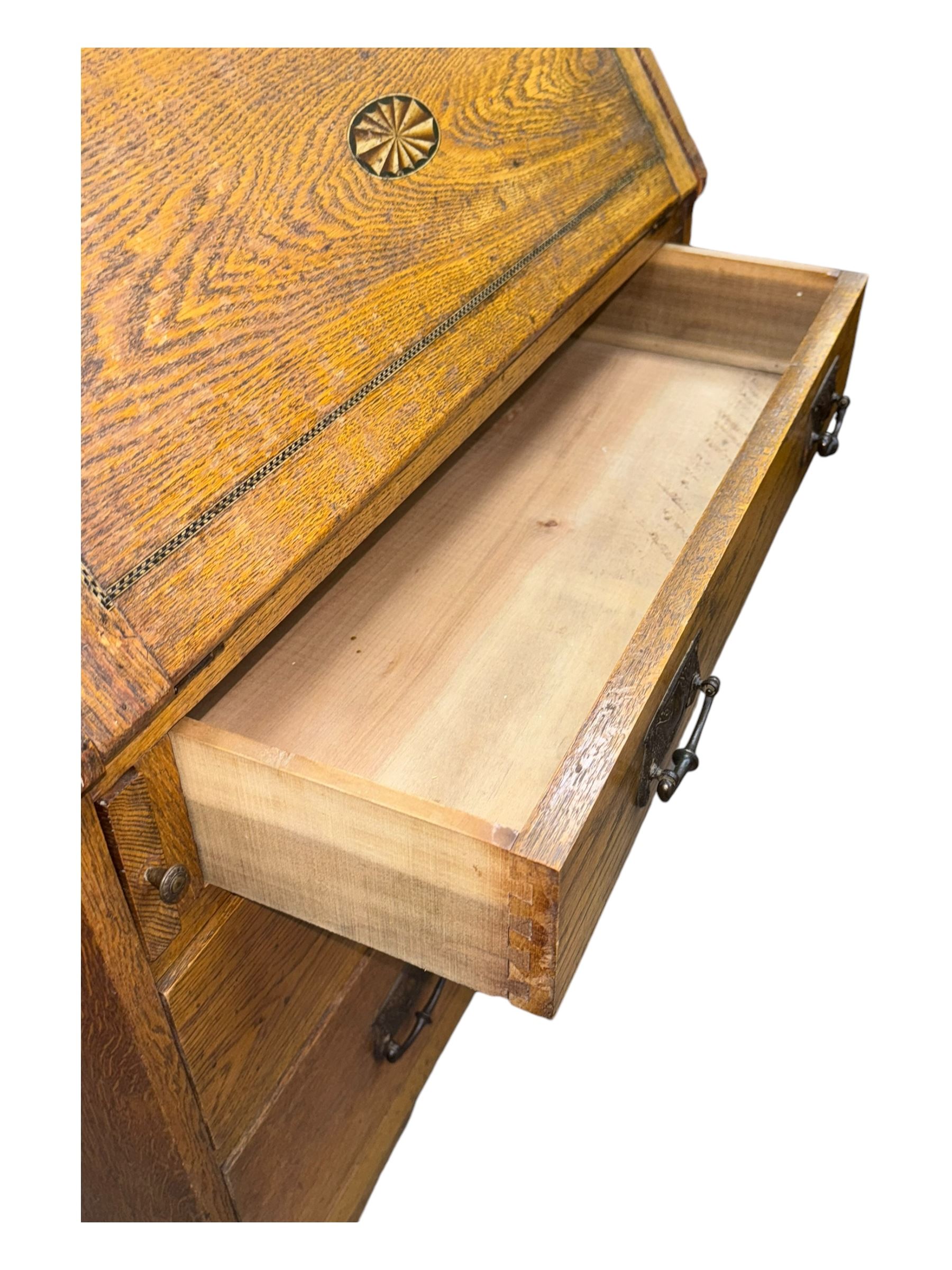Early 20th century Arts and Crafts oak bureau bookcase, twin leaded and glazed stained glass doors enclosing two shelves, over bureau section with fall-front inlaid with sunburst motif, fitted interior of pigeonholes and shelves, on bracket feet