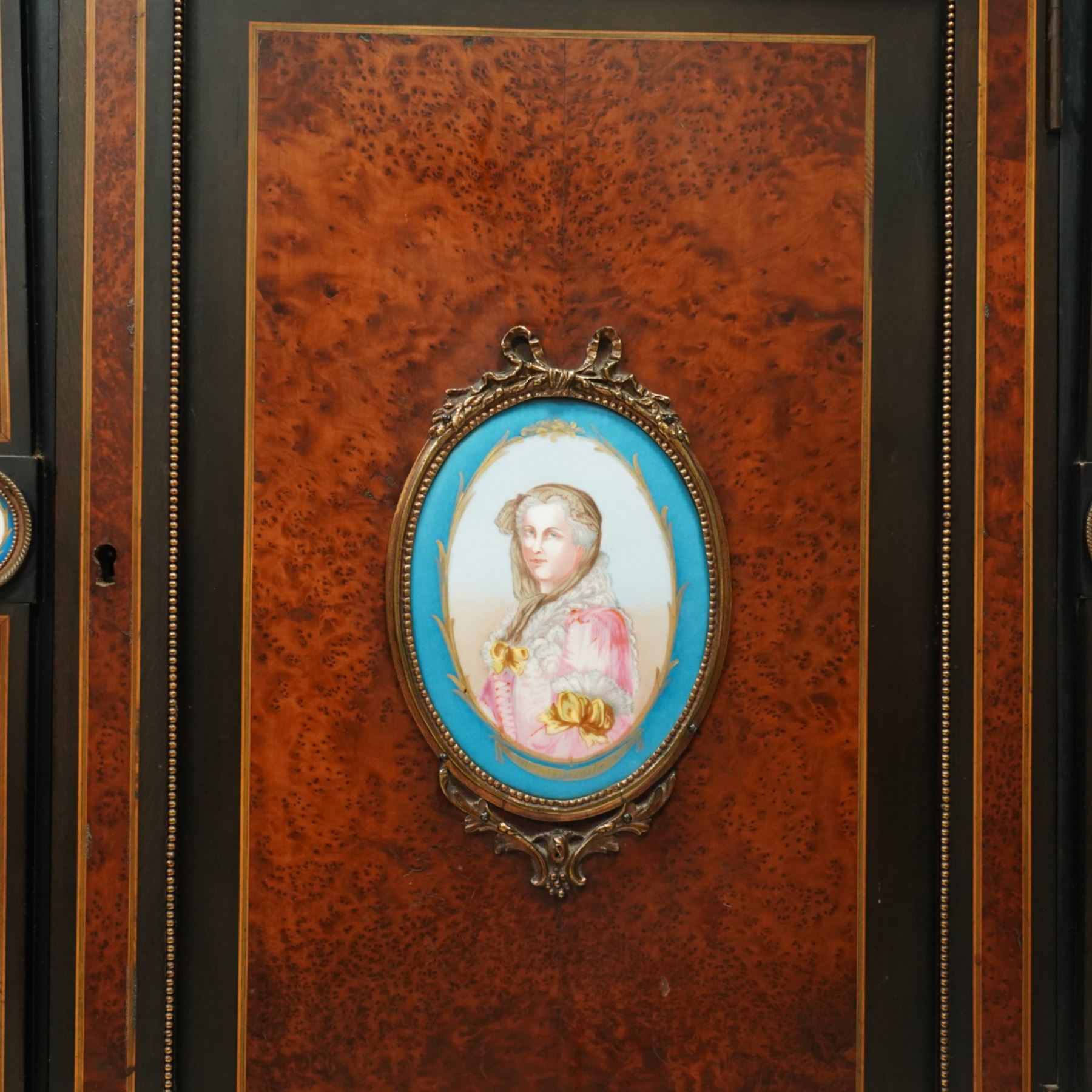 Victorian Aesthetic Movement amboyna and ebonised cabinet, the arched raised back inlaid with amboyna and floral porcelain plaque, over a raised shelf with fluted and turned supports, and a bevelled mirror plate with brass beading beneath, the inverted-breakfront top over an ornate frieze drawer with guilloche style gilt-metal mounts and porcelain plaques, the panelled door with a central porcelain plaque depicting a lady in a fuchsia gown within concentric ebonised and amboyna inlays, flanked by architectural style uprights with Corinthian capitals, raised on turned feet