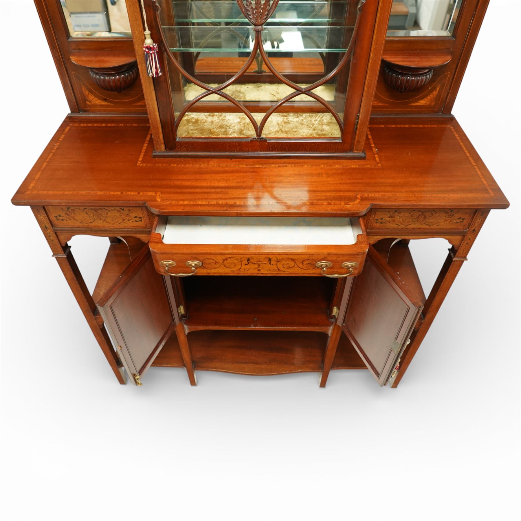 Edwardian Sheraton Revival inlaid mahogany breakfront cabinet, the broken swan neck pediment with central finial inlaid with cartouche and scrolling floral festoons, the central cabinet fitted with astragal-style glazed door with central urn and swag motif, flanked by arched bevelled mirrors over a shelf and gadrooned bracket over paterae inlays, base fitted with marquetry frieze rawer over a panelled cupboard with intricate inlaid scrolling floral designs, flanked by open shelves, raised on square tapering herringbone inlaid supports united by undertier
