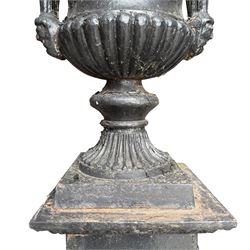 Pair of Victorian cast iron campana form garden urns, the gadrooned flared edge over a scrolling acanthus relief body, with twin male mask handles to each side, over a lobed underbelly with fluted socle, the rectangular pedestals with relief floral garlands and stepped base, in rustic black-painted finish, planted