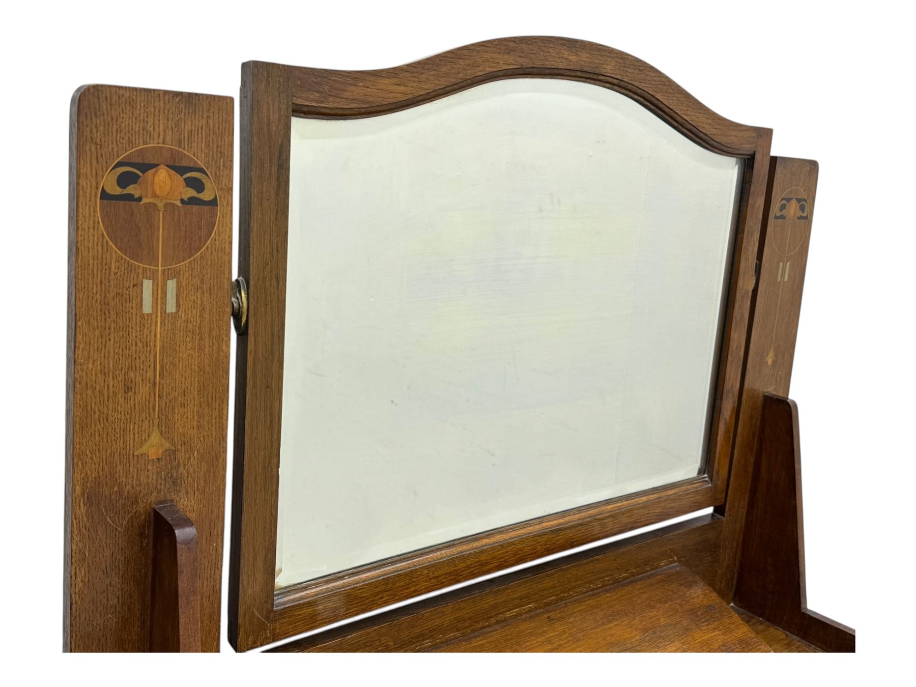 Early 20th century Arts and Crafts oak dressing chest, the arched mirror flanked by floral marquetry inlaid uprights above a shelf and drawer, over three long drawers with square hammered metal plates and circular pulls, on slender supports with castors