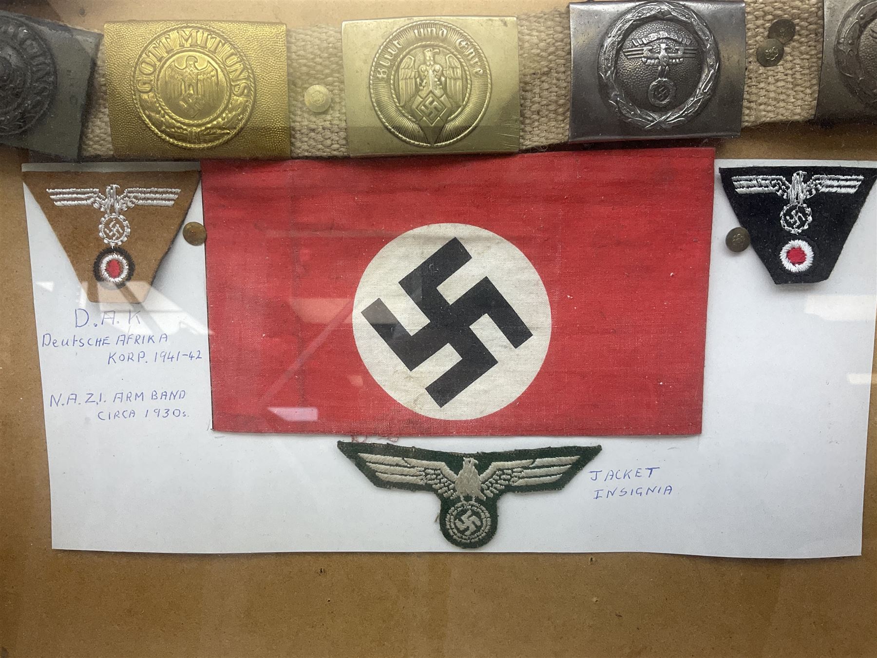 Display of eleven German belt buckles including 'SS', Party Buckle, two Army, Fire Service, two Luftwaffe, NSKK, Hitler Youth and WW1 Army; framed with three cloth badges and an armband