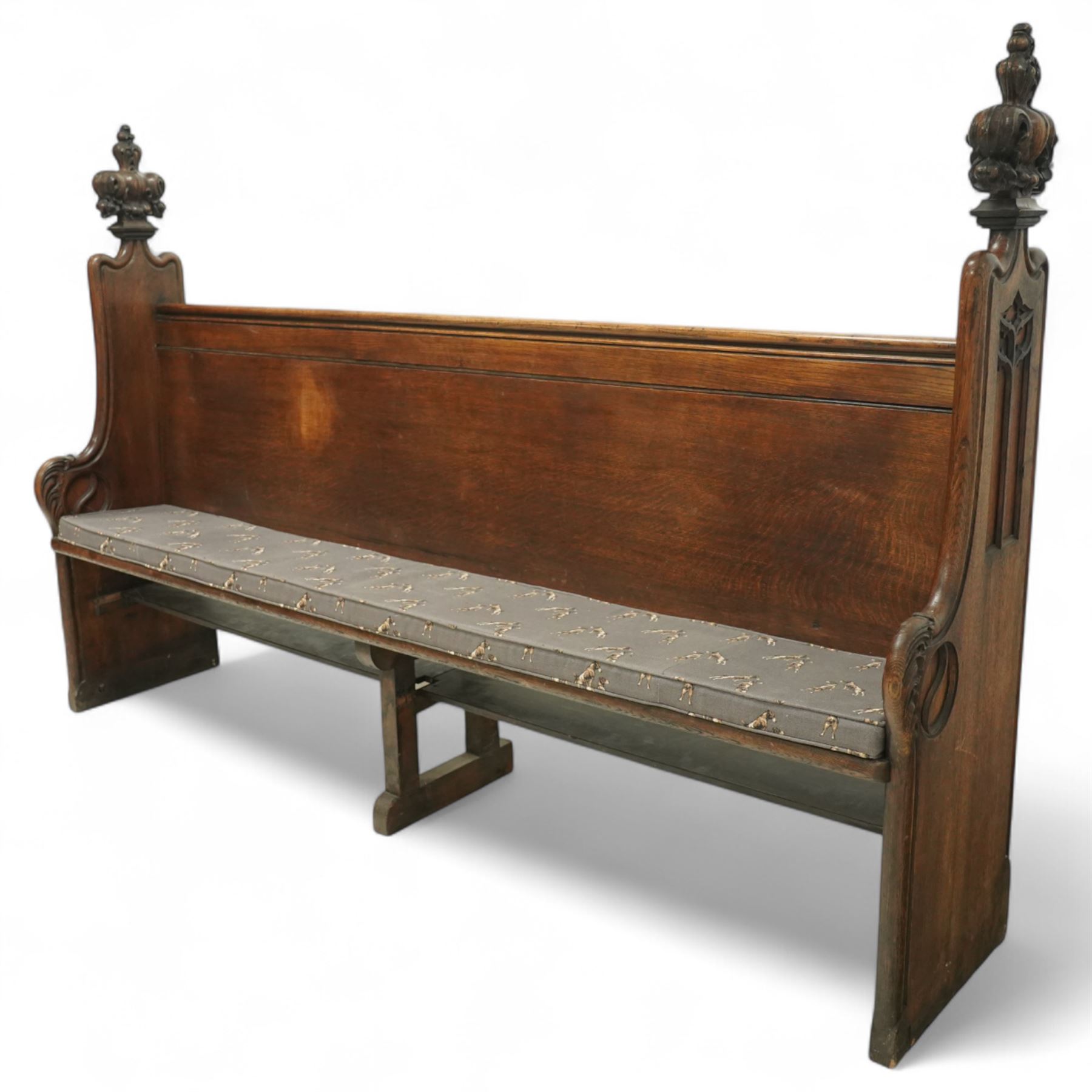 Late 19th century Gothic revival oak church pew, with high backrest framed by carved stylised cross panels and crown-topped finials, shaped end panels with scroll detailing, on block end supports and central stretcher, with later padded seat cushion