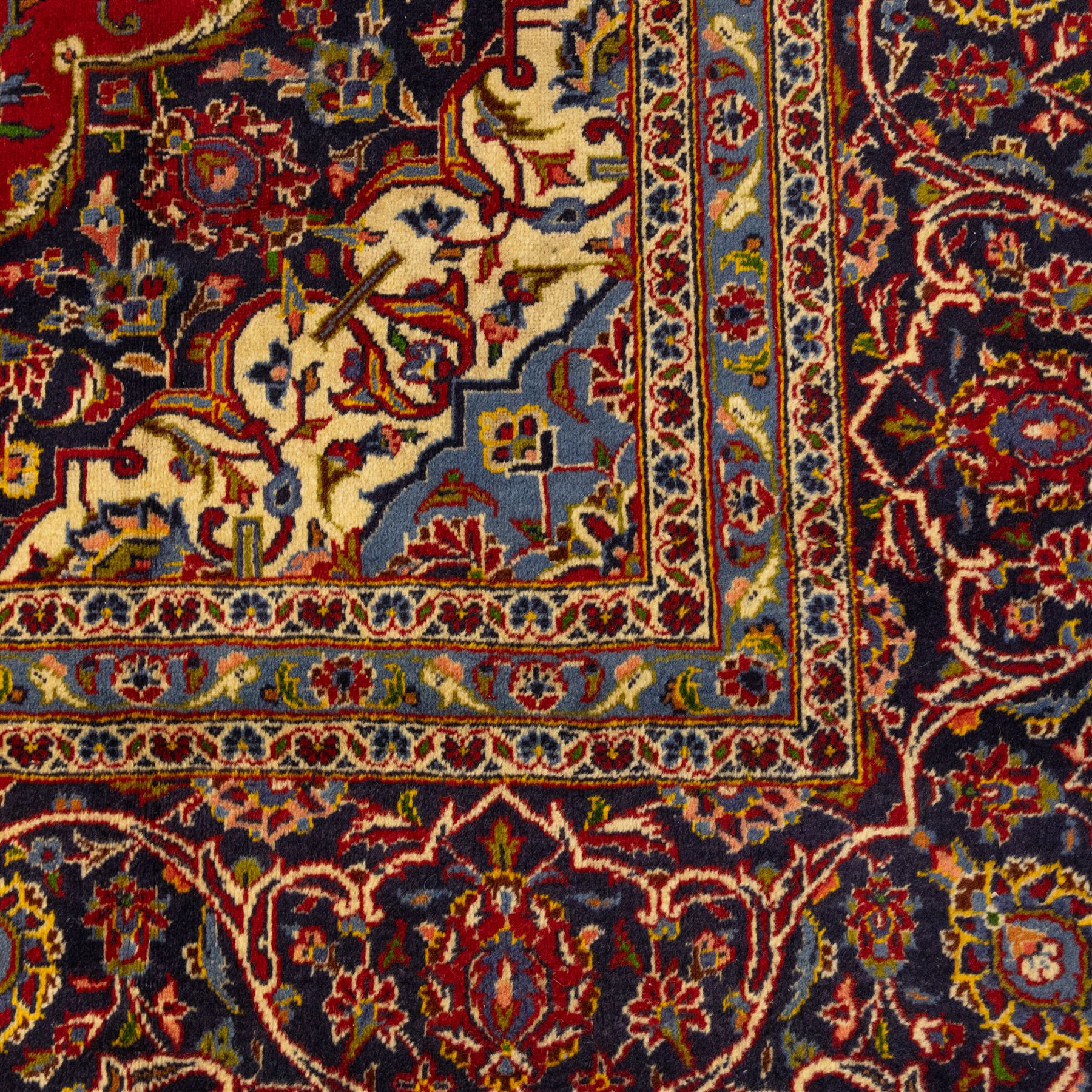 Central Persian Kashan hand-knotted carpet, crimson ground, with a central shaped palmette medallion with pendants, surrounded by scrolling foliate and floral motifs incorporating rosettes, palmettes, and vine work, enclosed by a dark navy with repeating border with repeating palmettes and meandering vines, flanked by floral guard stripes, with cotton fringes 