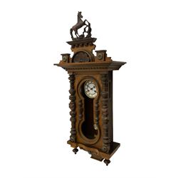 A late 19th century German wall clock in a mahogany case with a decorative carved pediment, full length shaped glazed door (glass missing) flanked by turned columns and pendant finials, with a two part enamel dial and steel gothic hands, eight-day striking movement striking the hours and half hours on a coiled gong, with a gridiron R/A pendulum. 

