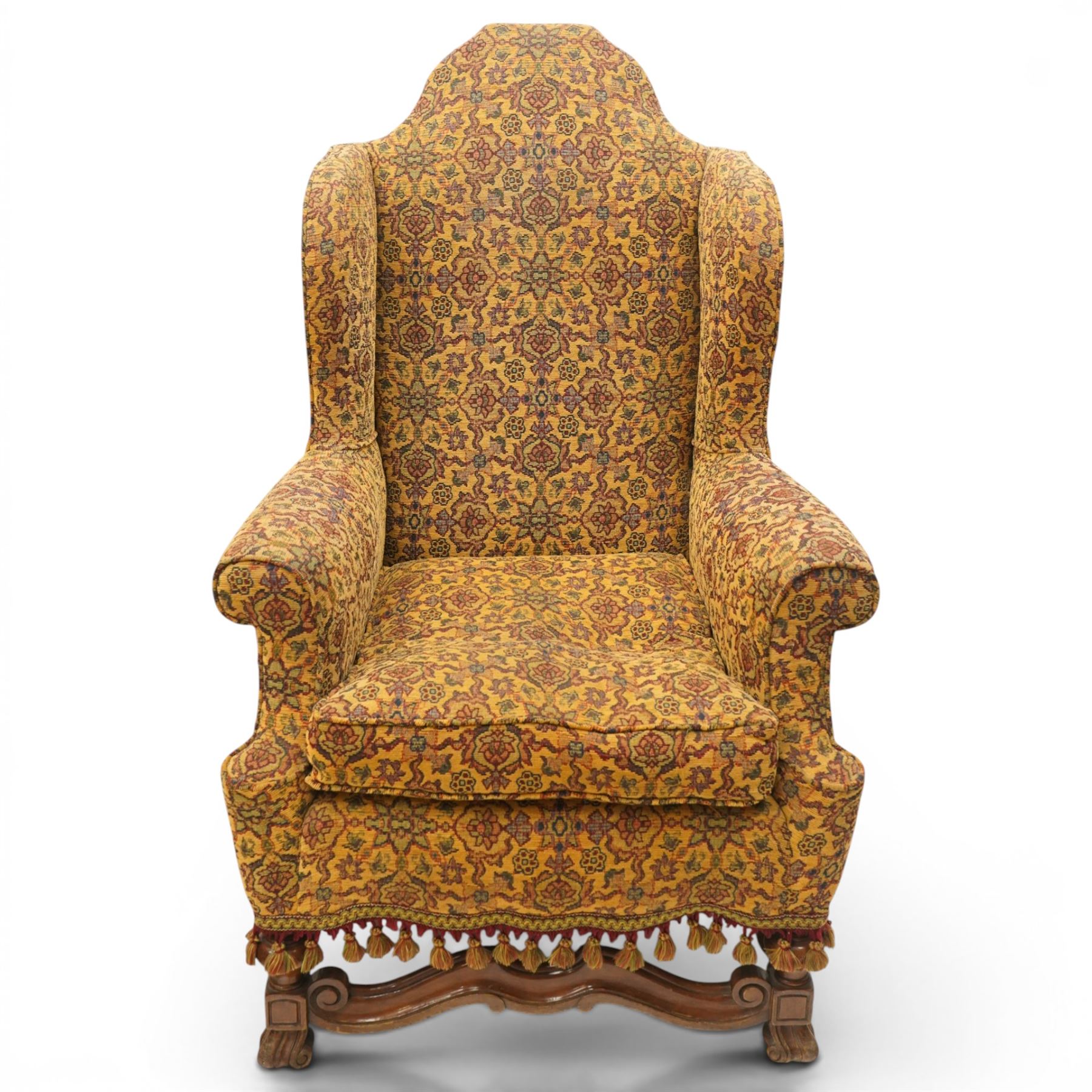 Pair of William & Mary design wingback armchairs, the arched high-back over rolled arms with a loose seat cushion and sprung seat, upholstered in old-gold ground foliate patterned fabric, raise on scroll feet united by a front serpentine stretcher