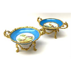 A pair of early 20th century Sevres style ormolu mounted twin handled tazzas, painted with central panels of exotic birds within a blue border, one example with spurious serves mark beneath, H11cm, L21cm. 