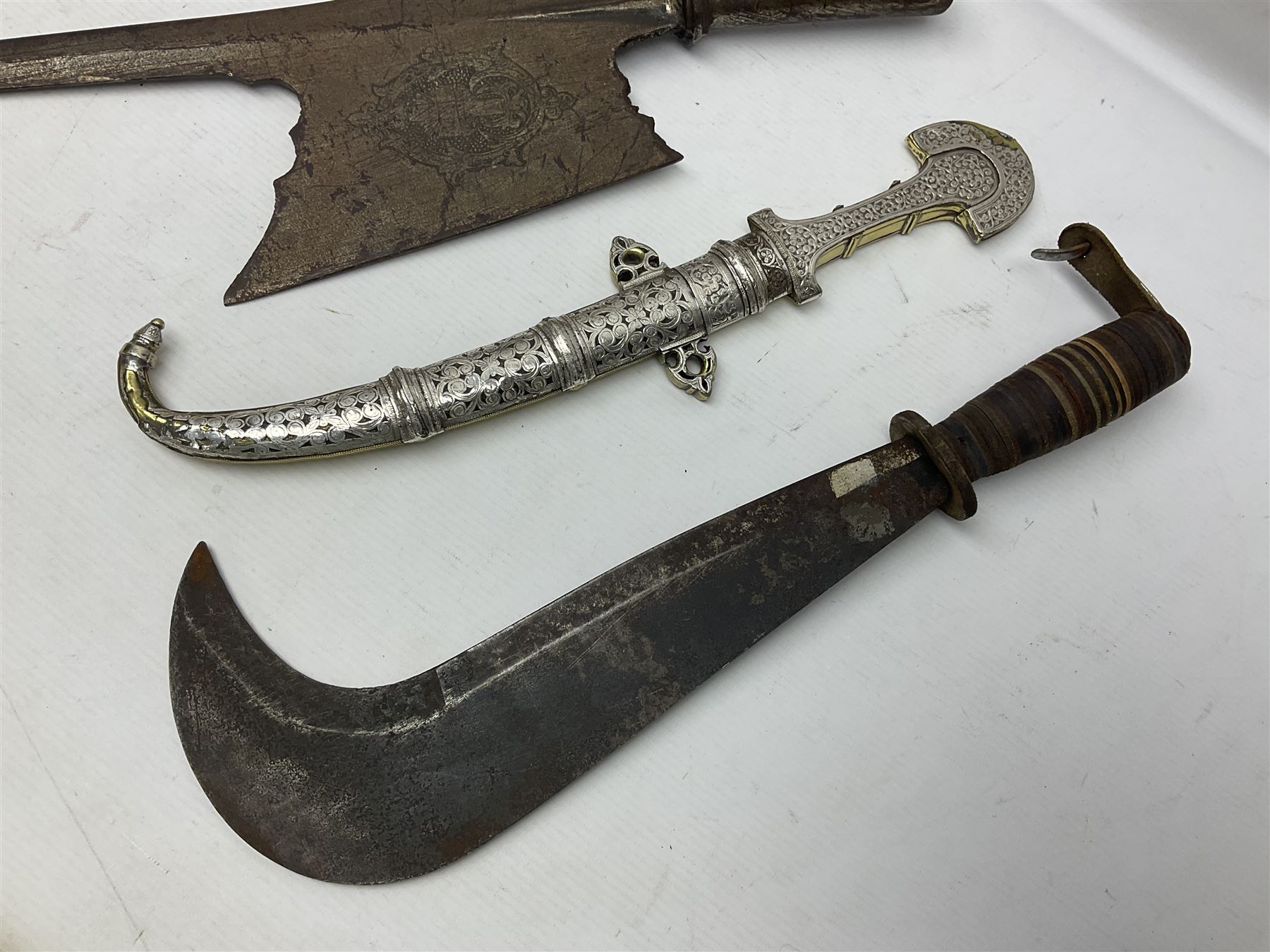 Moroccan jambiya dagger with 24.5cm plain curved blade stamped LONDON to the ricasso area, the hilt and scabbard overlaid with plain sheet brass to the back and ornate silver coloured metal to the front; waisted grip and arched pommel; two pyramid mounts to the scabbard L41cm overall; large billhook with curving blade and stacked leather washer grip; and a reproduction halberd blade (3)