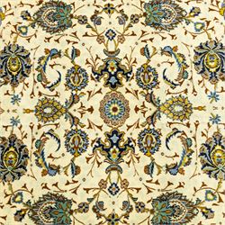 Central Persian Kashan hand-knotted carpet, ivory ground, with an all-over floral design of palmettes, scrolling vines and flowerheads in blue, ochre and beige tones, enclosed by a sage green border with repeating floral sprays and leafy scrolls, flanked by narrow guard stripes, with cotton fringes