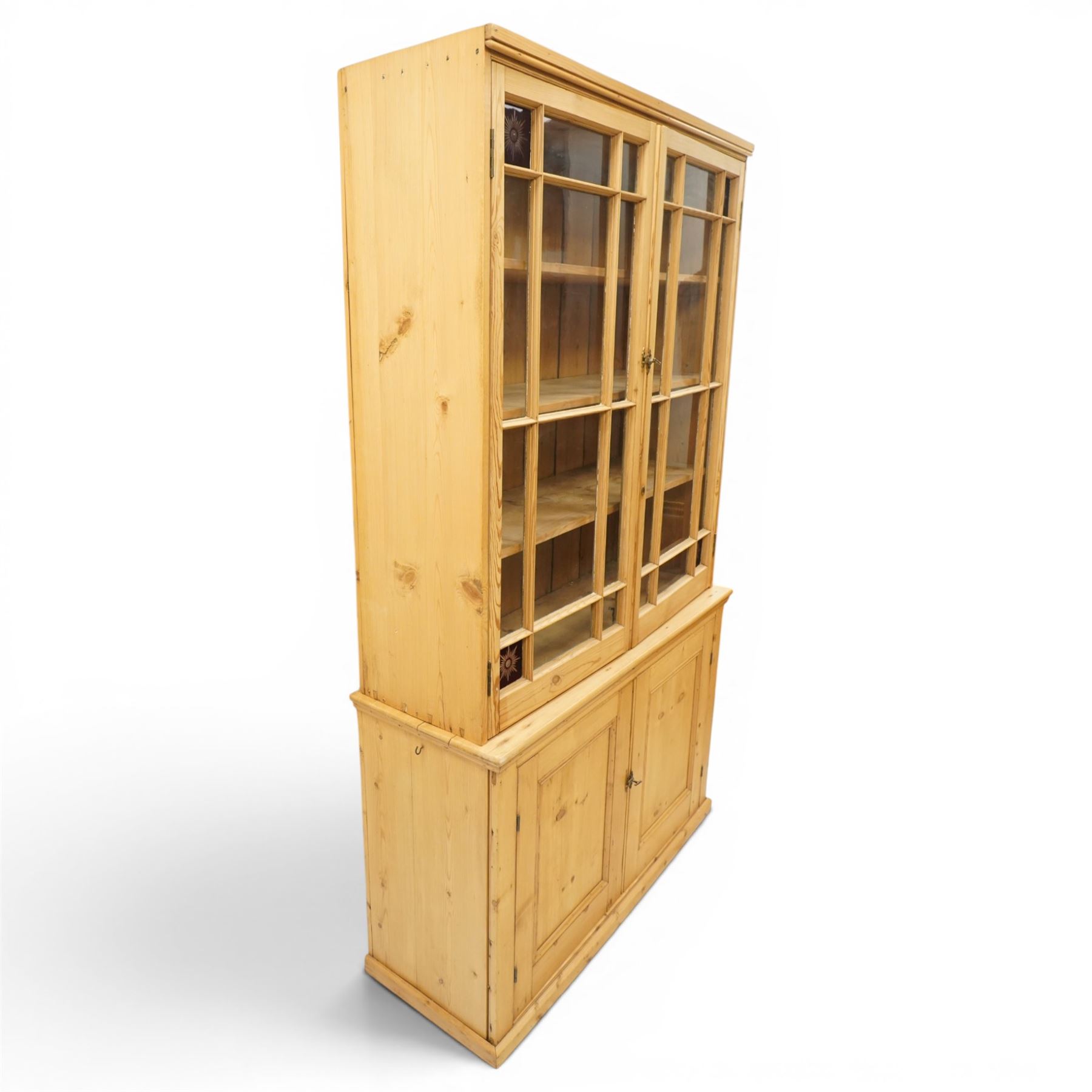 19th century pine glazed bookcase cupboard, twin astragal glazed doors with stained glass corner panels, enclosing fixed shelves, the lower section fitted with two panelled cupboard doors enclosing single shelf, on plinth base