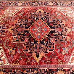 North West Persian Heriz hand-knotted carpet, madder red field with large central stepped medallion with angular floral and geometric designs, surrounded by stylized palmettes, leaves and tribal motifs with indigo corner spandrels, enclosed by a broad navy border of geometric floral devices between multiple guard stripes, with cotton fringe ends 
