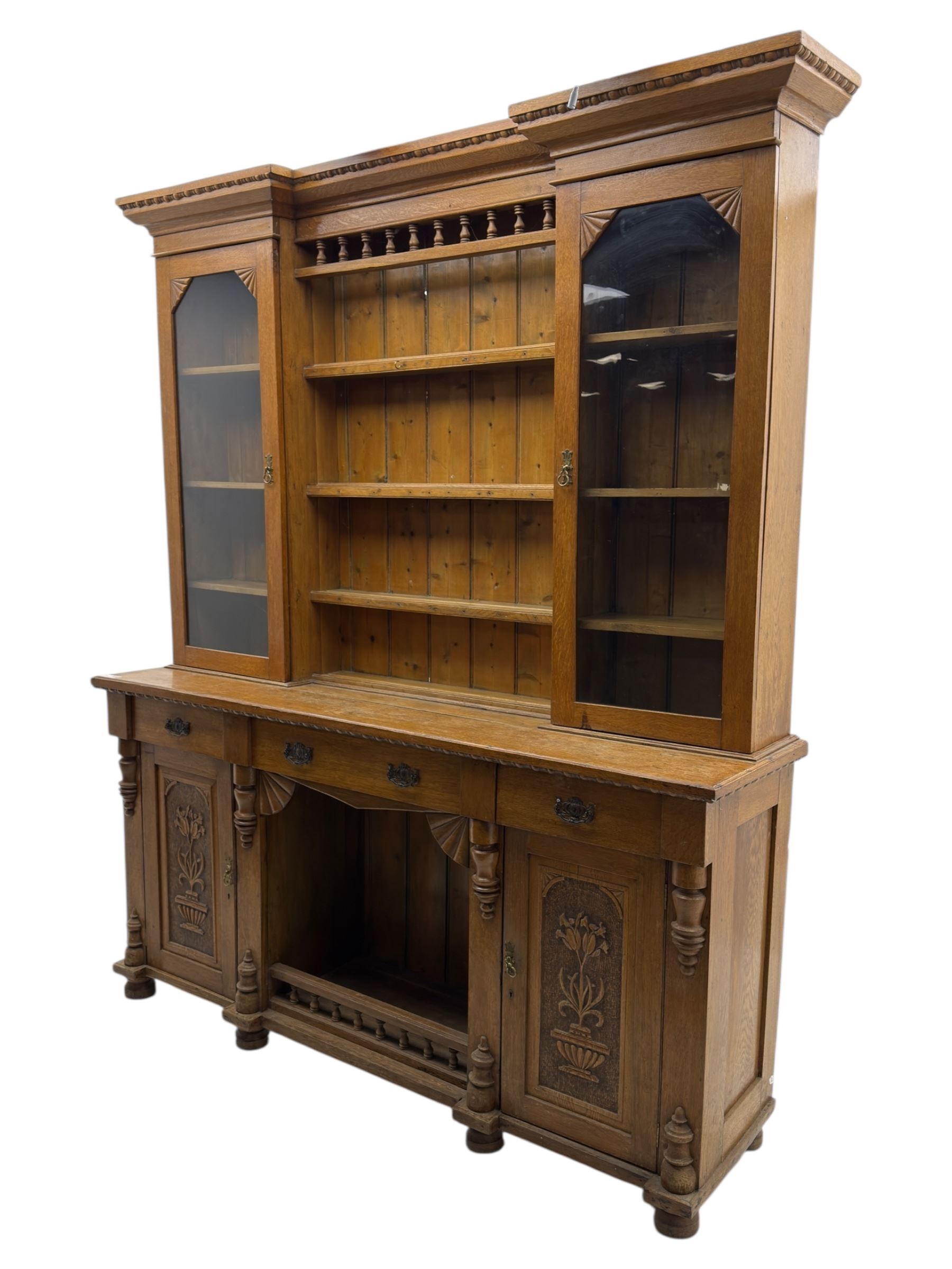 Late 19th century pine dresser, the projecting cornice with dentil and spindle moulded frieze above a three-tier plate shelves, flanked by glazed doors, the base fitted with three frieze drawers over arched apron and carved floral panelled cupboard doors, raised on turned feet