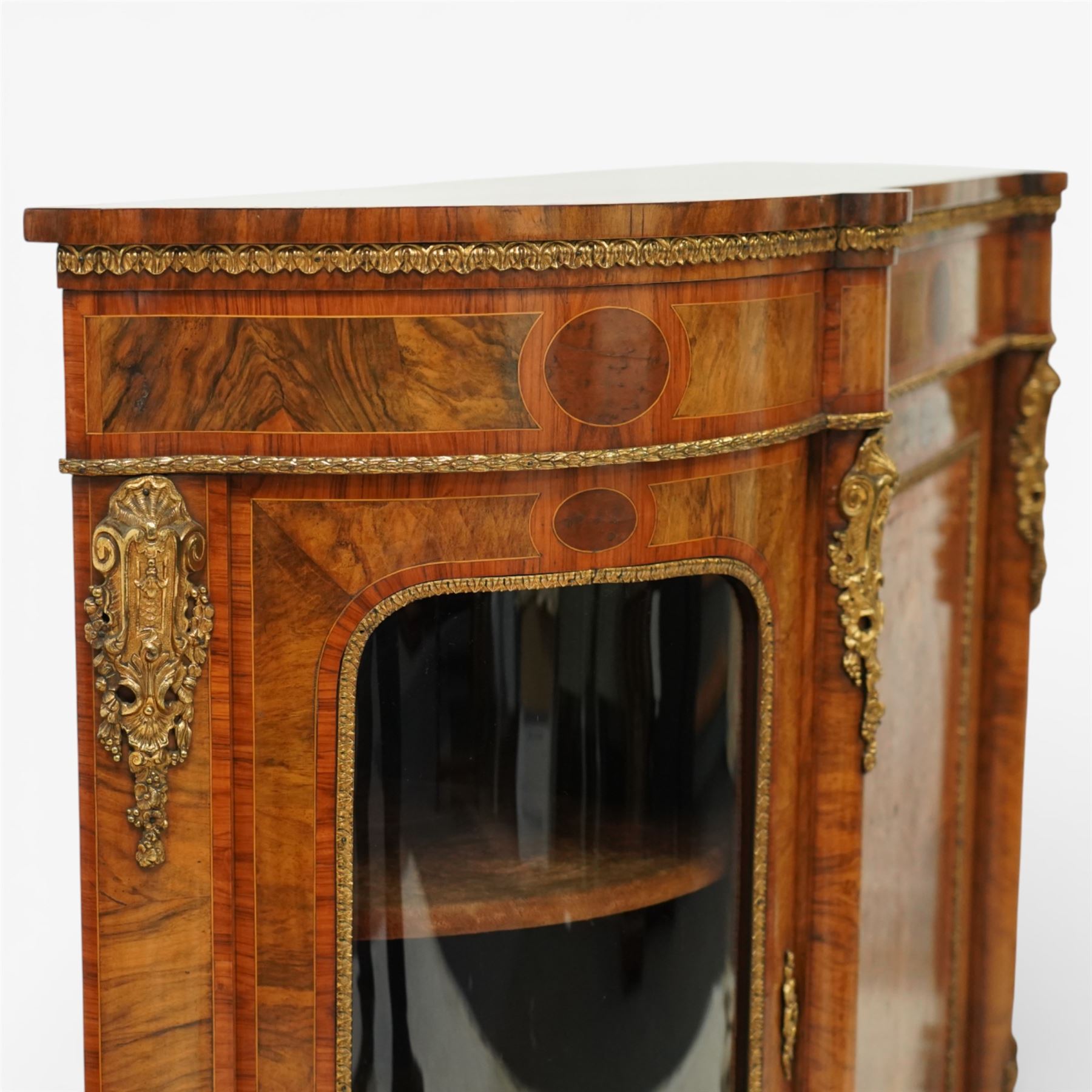 Late Victorian figured walnut credenza, the shaped demi-lune top with cross-banding and stringing over an inlaid frieze with foliate gilt-metal mounts, fitted with a central geometric-inlaid panelled door, flanked by convex-glazed side-cabinets with a gilt metal slip and outer-banding, the uprights surmounted by gilt-metal cartouche mounts with cascading foliage details, raised on a shaped skirted base with central female-mask gilt mount