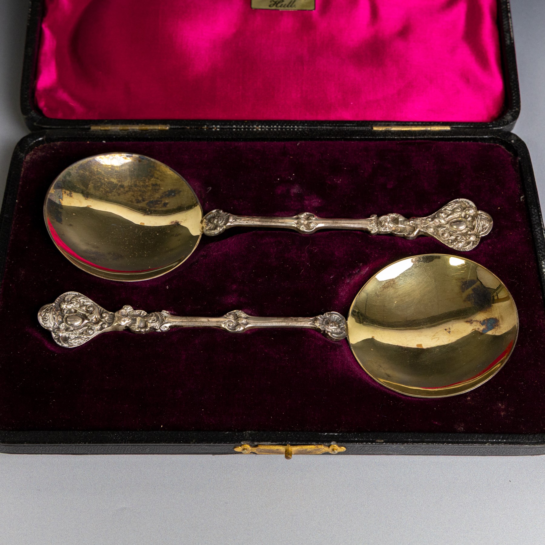 Pair of late Victorian silver serving spoons, each with circular gilt bowls and embossed putti decoration, hallmarked William Hutton & Sons Ltd, London 1895, contained within a tooled leather box with burgundy velvet and silk interior