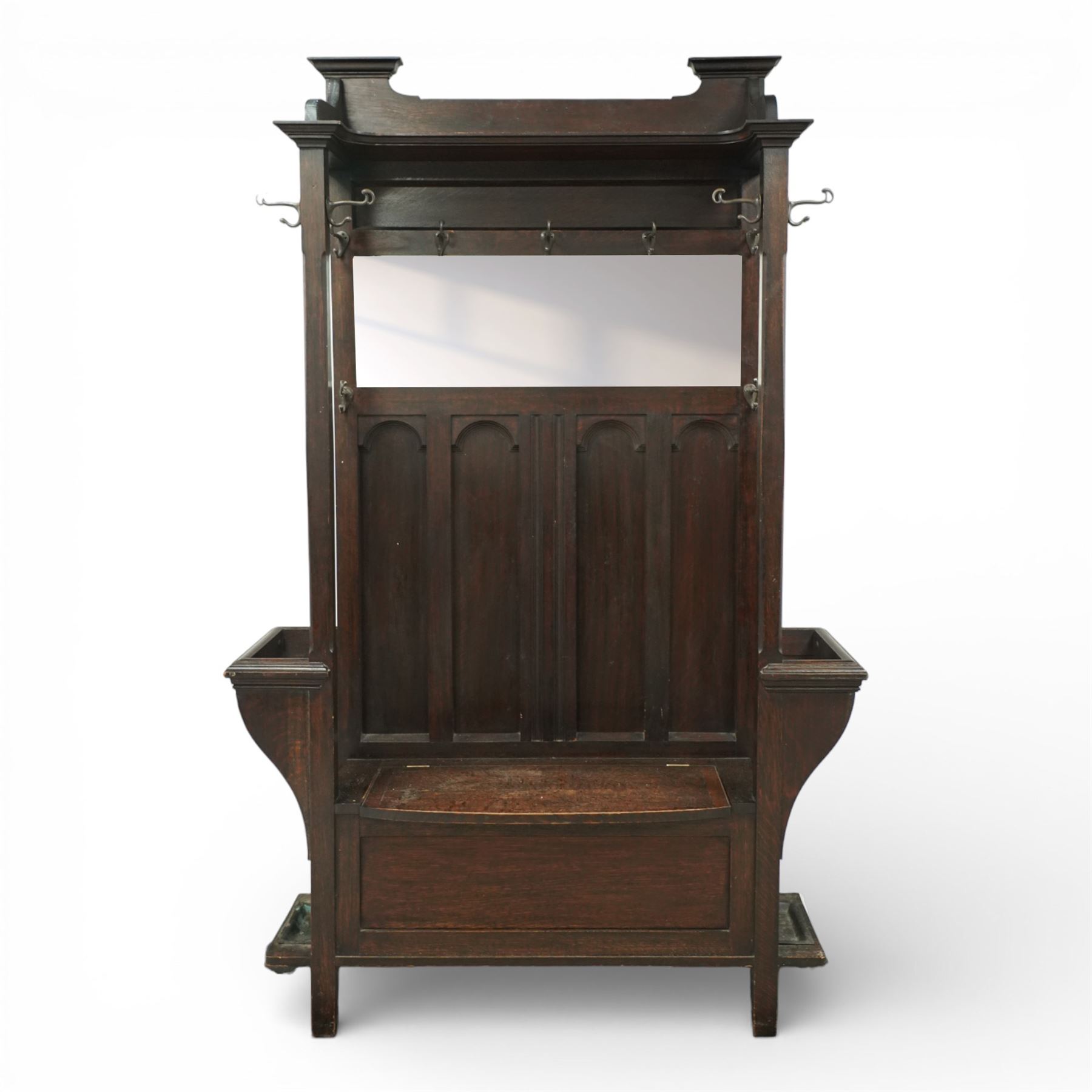 Arts and Crafts design oak settle, with projecting moulded cornice over a boarded back fitted with a rectangular bevelled mirror plate and a row of wrought iron coat hooks, the panelled back with arched fielded panels, flanked by shaped umbrella or stick stands with removable drip trays, the plank hinged seat over a panelled box base, on square section supports