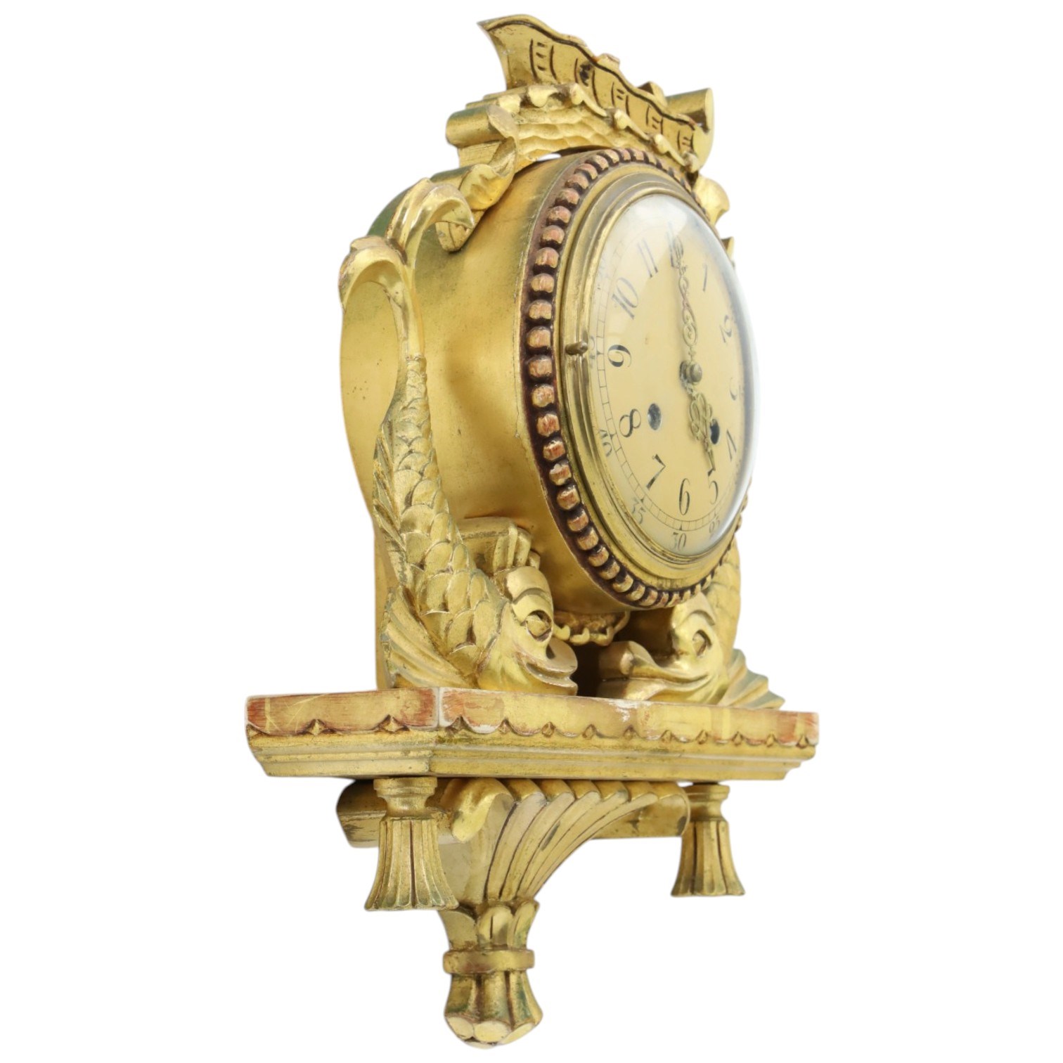 20th century Continental giltwood wall clock, circular dial with Arabic numerals and pierced gilt hands, within a carved case surmounted by a scrolling crest and flanked by stylised dolphins, above a shaped shelf with tassel pendants and a foliate terminal, pendulum missing