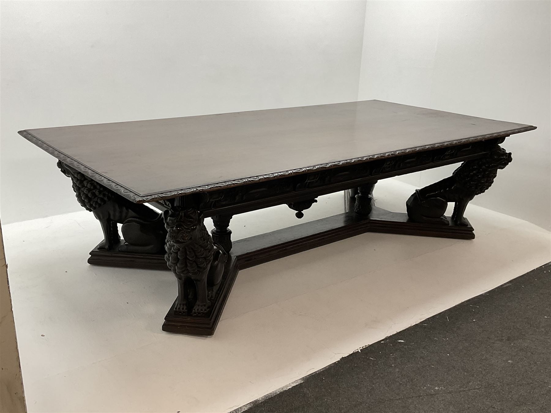 Large 19th century carved oak library table, the rectangular top with lunette carved edge over cartouche panelled and carved  scroll frieze, the central tapering rectangular support relief carved with masks and garlands, on four carved seated lion supports at each corner, on flat angular stretcher with recessed castors stamped ‘Bartholomew & Fletcher, London’