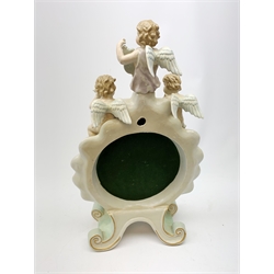  A late 19th century Sitzendorf porcelain mirror, the mirror plate of circular form set within a shaped surround encrusted with flowers and foliage, surmounted by three figures of cherubs and raised upon four scroll feet, with blue cross hatch mark beneath, H57cm.   