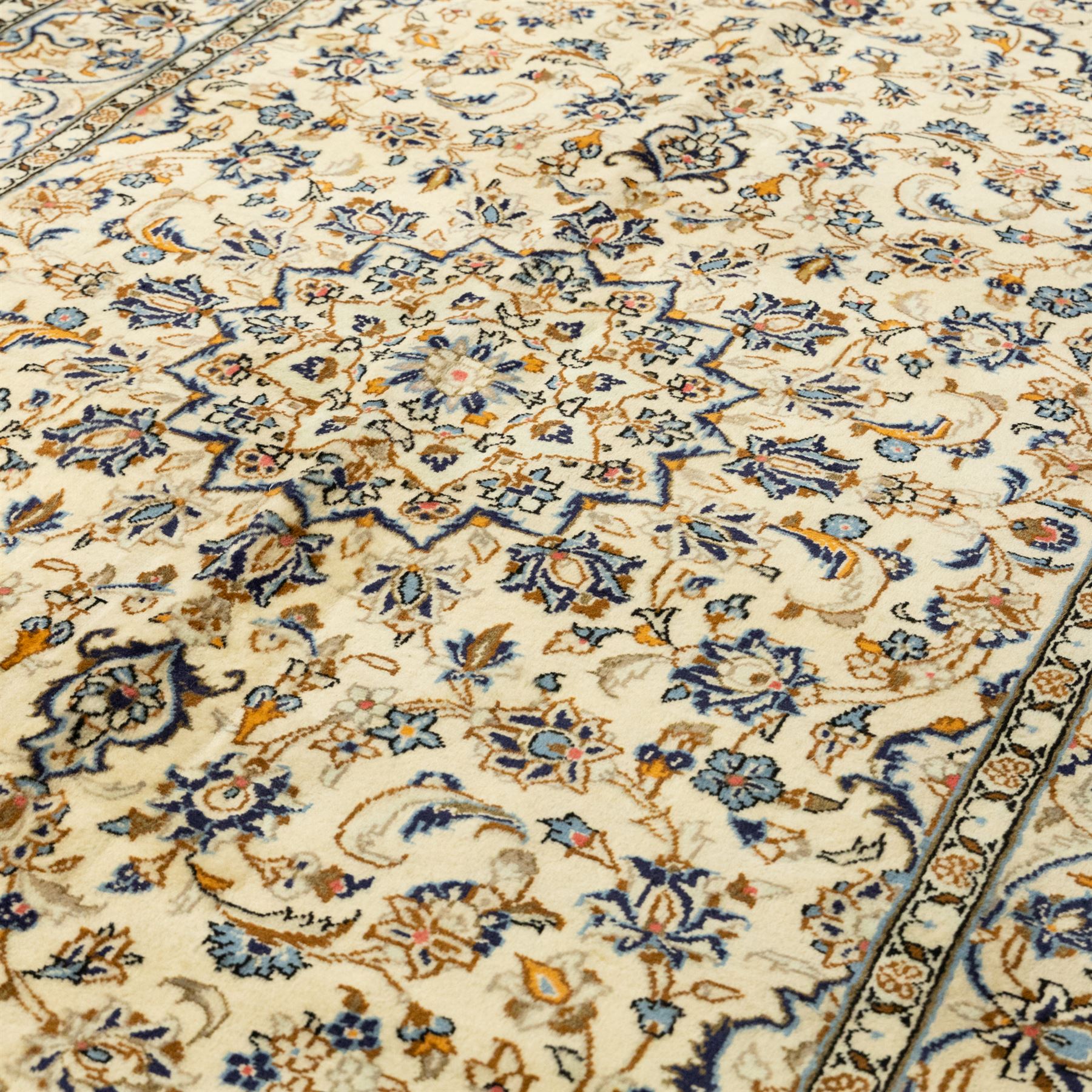 Central Persian Kashan hand-knotted rug, ivory ground, with a small central floral medallion and pendants, the field decorated with fine blue, ochre and beige scrolling vines, flowerheads and palmettes, within an ivory border of repeating floral sprays and meandering vines, flanked by narrow guard stripes, with cotton fringes