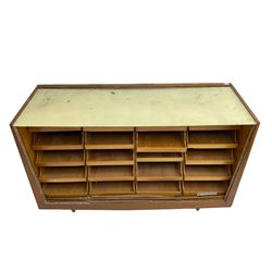 Mid-20th century teak haberdashery shop cabinet, sloped front and fitted with twenty drawers, metal cup handles and label holders, glass top with bass rule, on turned tapering feet