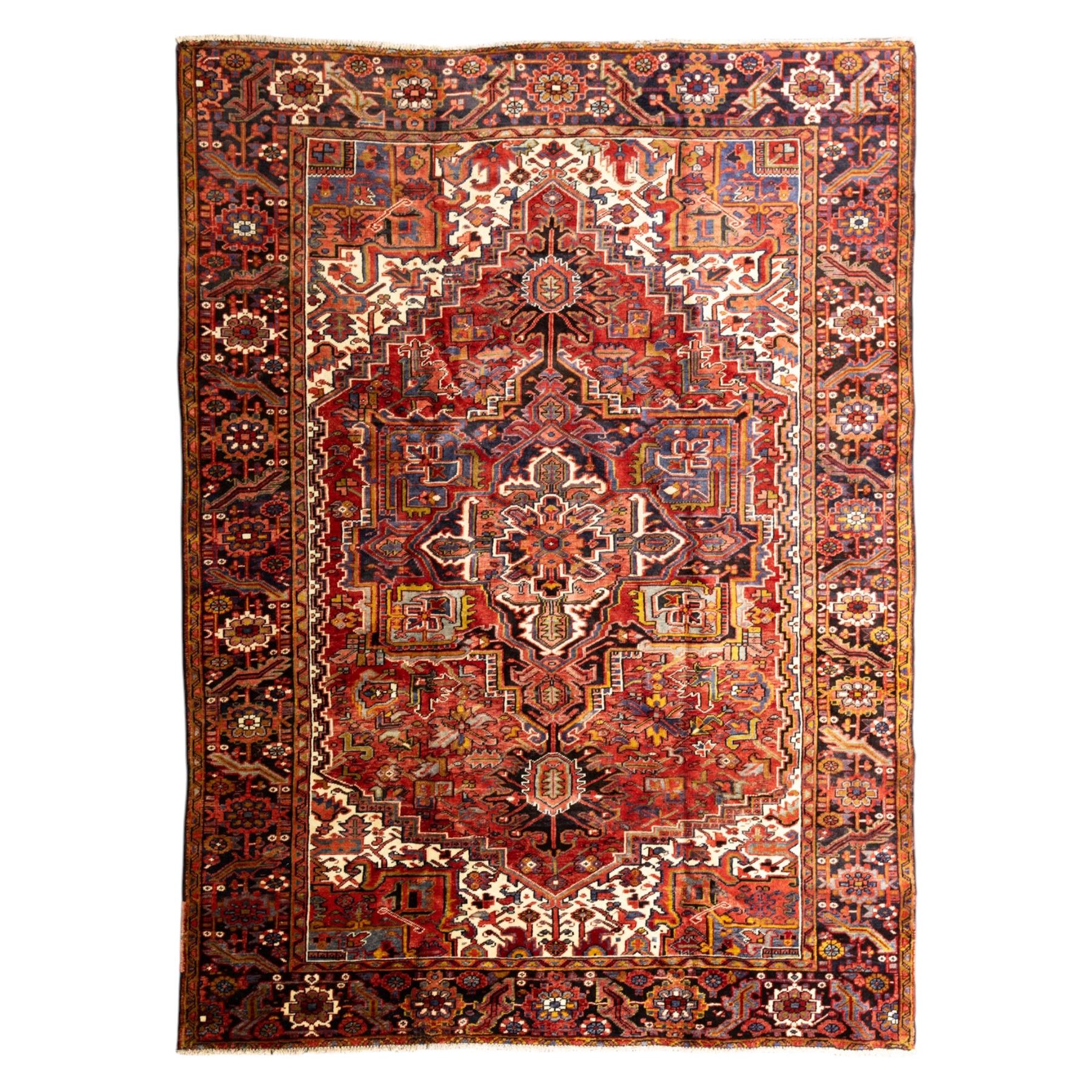 North West Persian Heriz hand-knotted carpet, madder red ground, with a large central geometric medallion with pendants, surrounded by angular foliate and floral motifs incorporating stylised rosettes, palmettes and serrated vine work, enclosed by ivory spandrels and a dark navy border with repeating geometric flowerheads and angular meandering vines, flanked by narrow patterned guard stripes, with cotton fringes