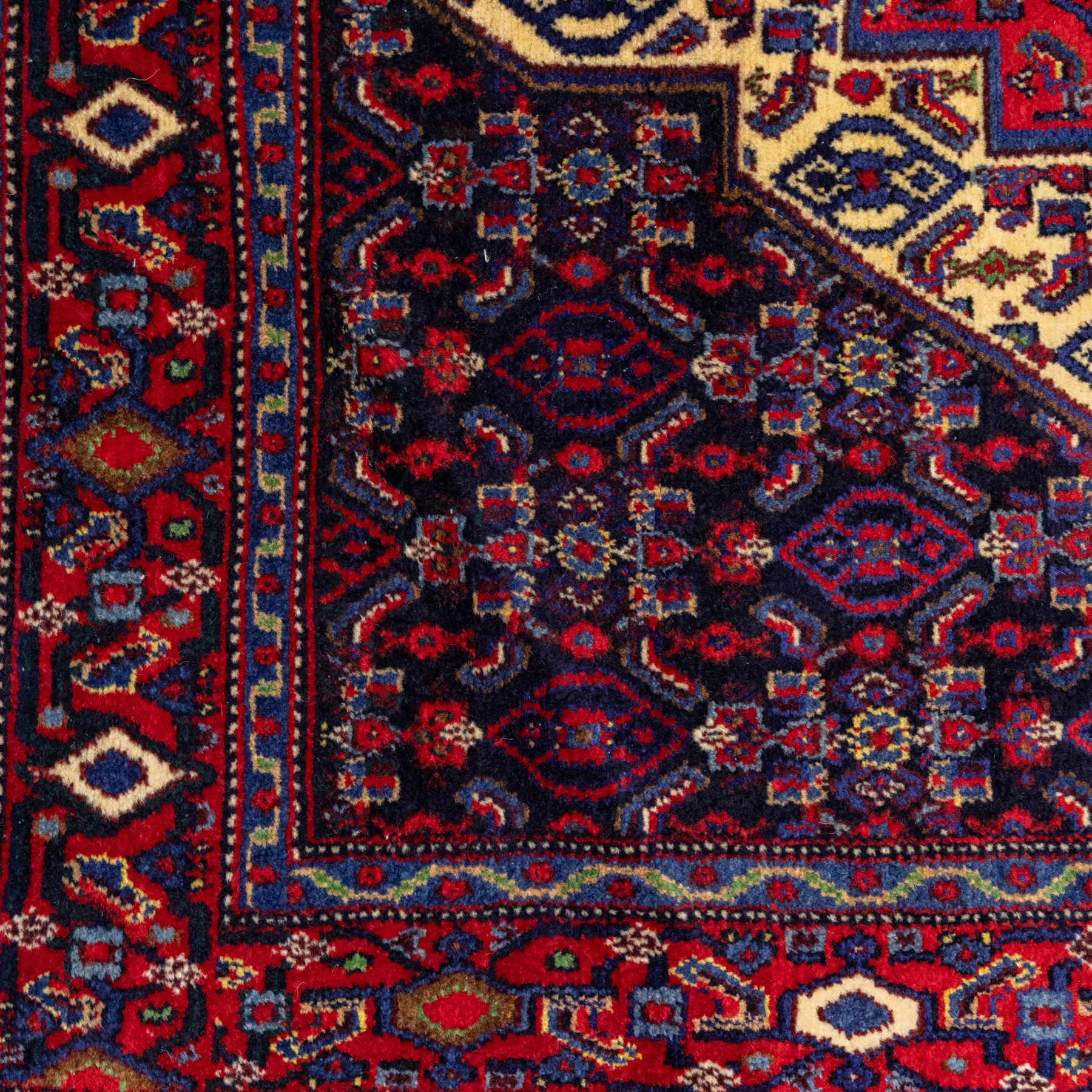 North West Persian Senneh hand-knotted rug, dark indigo field with a central bold stepped lozenge medallion of madder red and deep blue, enclosed within an ivory serrated contour forming a cruciform outline, medallion with repeating boteh and floral devices, field decorated with an all-over Herati pattern of rosettes enclosed by angular leaves principal border of madder red ground decorated with alternating floral heads, flanked by narrow guard stripes, with cotton fringe ends 