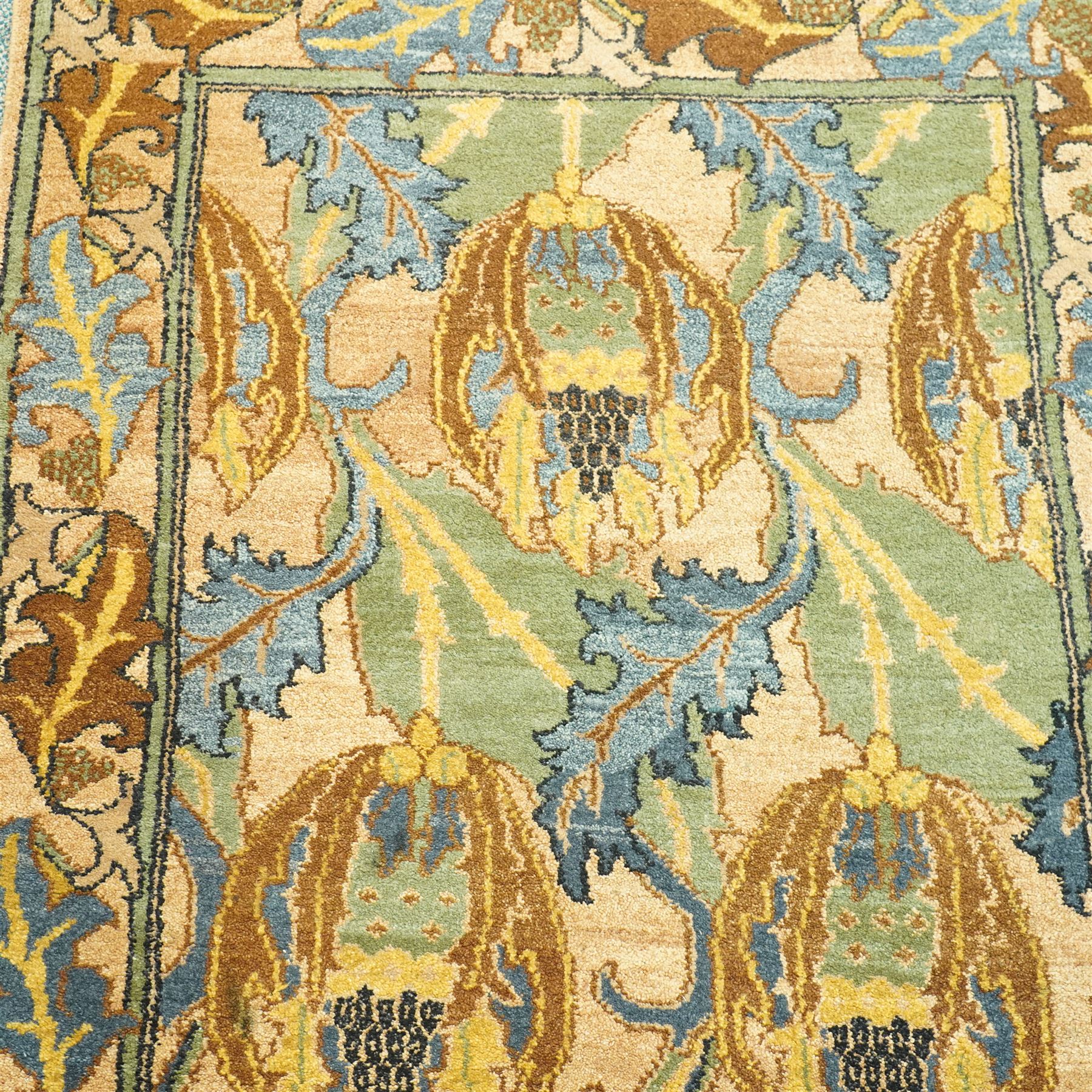 Arts & Crafts design wool pile rug, decorated with repeating stylized floral seed-head motifs and scrolling foliage within a complementary floral border in a muted palette of green, ochre, blue, and brown, with cotton fringe ends, with label for London House Oriental Rugs & Carpets  