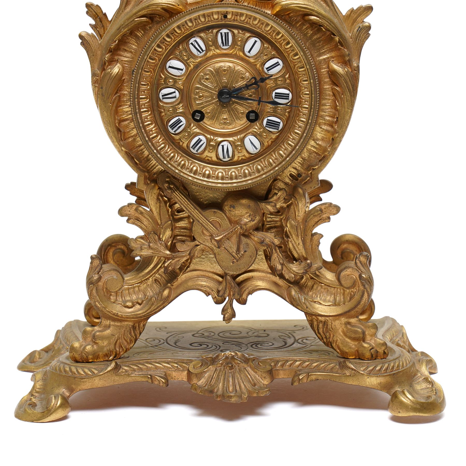 French-Late 19th century 8-day gilt brass rococo style mantel clock, c1890, waisted case  decorated with scroll work, leaves and musical instruments, resting on an attached plinth with splayed feet, dial within a glazed brass bezel with a gilt repoussé centre, porcelain cartouche Roman numerals and steel fleur-di-Lis hands, with a twin train movement, recoil anchor escapement and countwheel strike, sounding the hours and half-hours on a bell. With a matching pair of five-light candelabra. No Pendulum or Key.