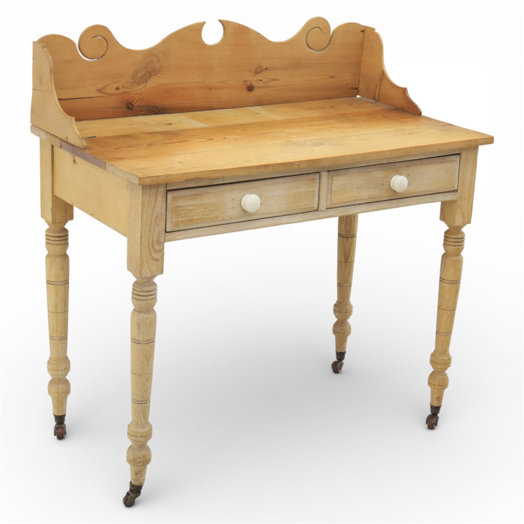 19th century stripped pine side table, the rectangular plank top with low shaped back rail incorporating scroll cut ends, fitted with two frieze drawers with ceramic knob handles, raised on turned supports terminating in castors