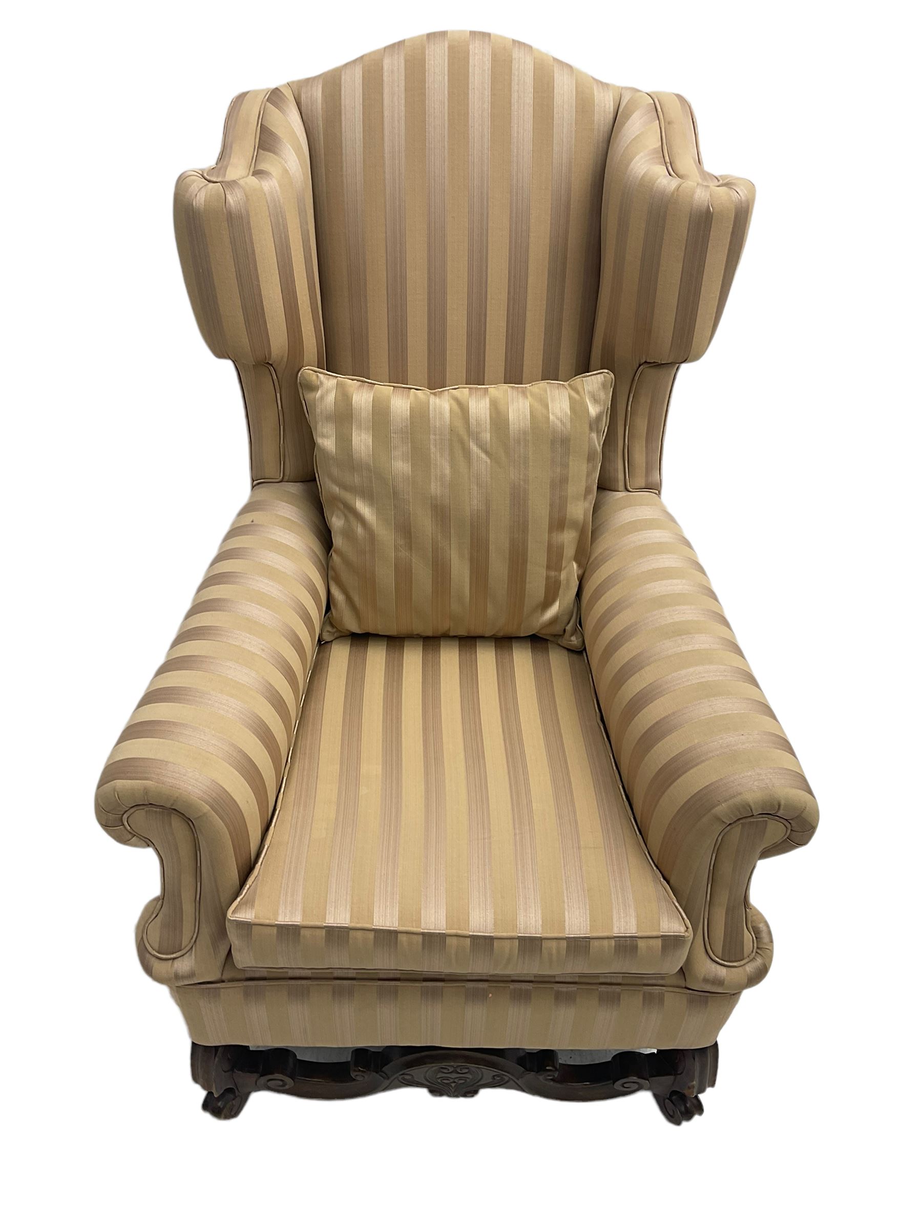 William and Mary style wingback armchair, upholstered in gold striped fabric, on walnut scroll carved supports joined by scroll and foliate carved middle rail, turned H-shaped stretchers, on castors
