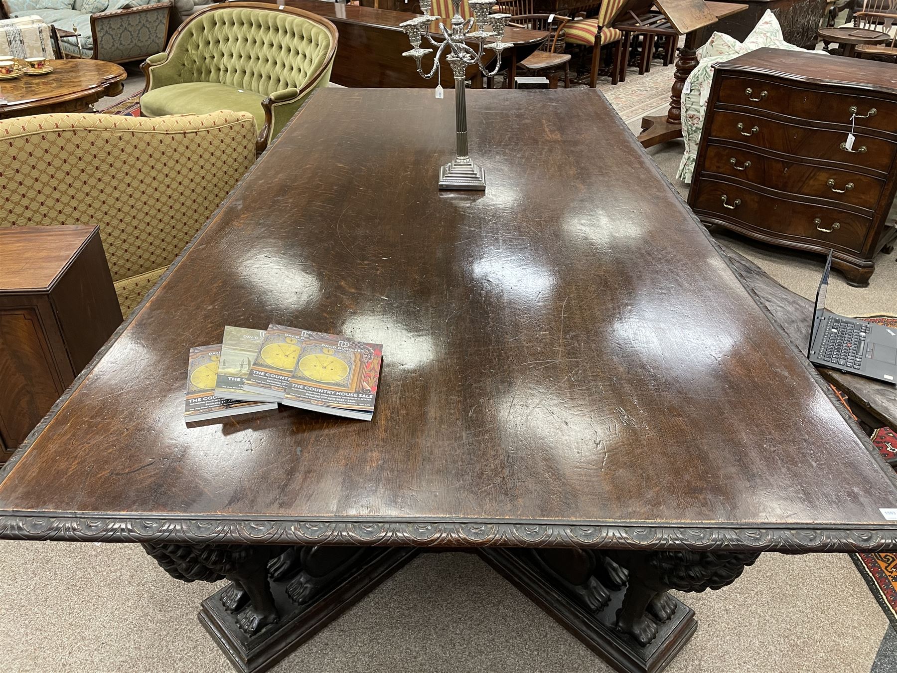 Large 19th century carved oak library table, the rectangular top with lunette carved edge over cartouche panelled and carved  scroll frieze, the central tapering rectangular support relief carved with masks and garlands, on four carved seated lion supports at each corner, on flat angular stretcher with recessed castors stamped ‘Bartholomew & Fletcher, London’