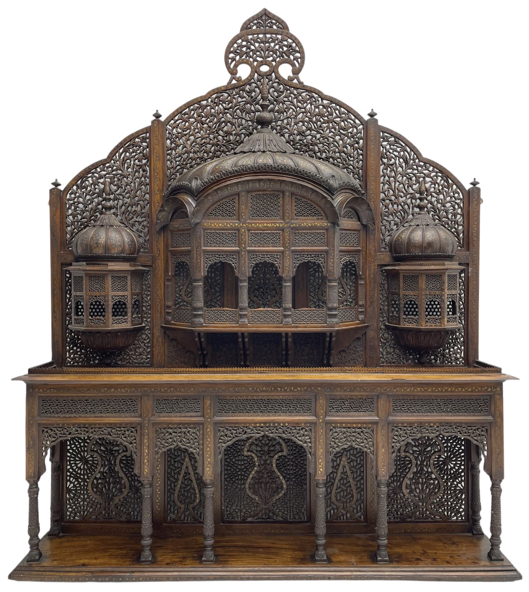 Late 19th century Indian Burmese carved and pierced hardwood and brass inlaid wall shelf, the shaped back profusely decorated with trailing and interlaced foliage, with projecting architectural structures, the central balcony with canopy top and five open windows with arched apertures on columns, two flanking five-sided balconies, the shelf supported by six foliate carved columns with pierced cusped pointed arches 