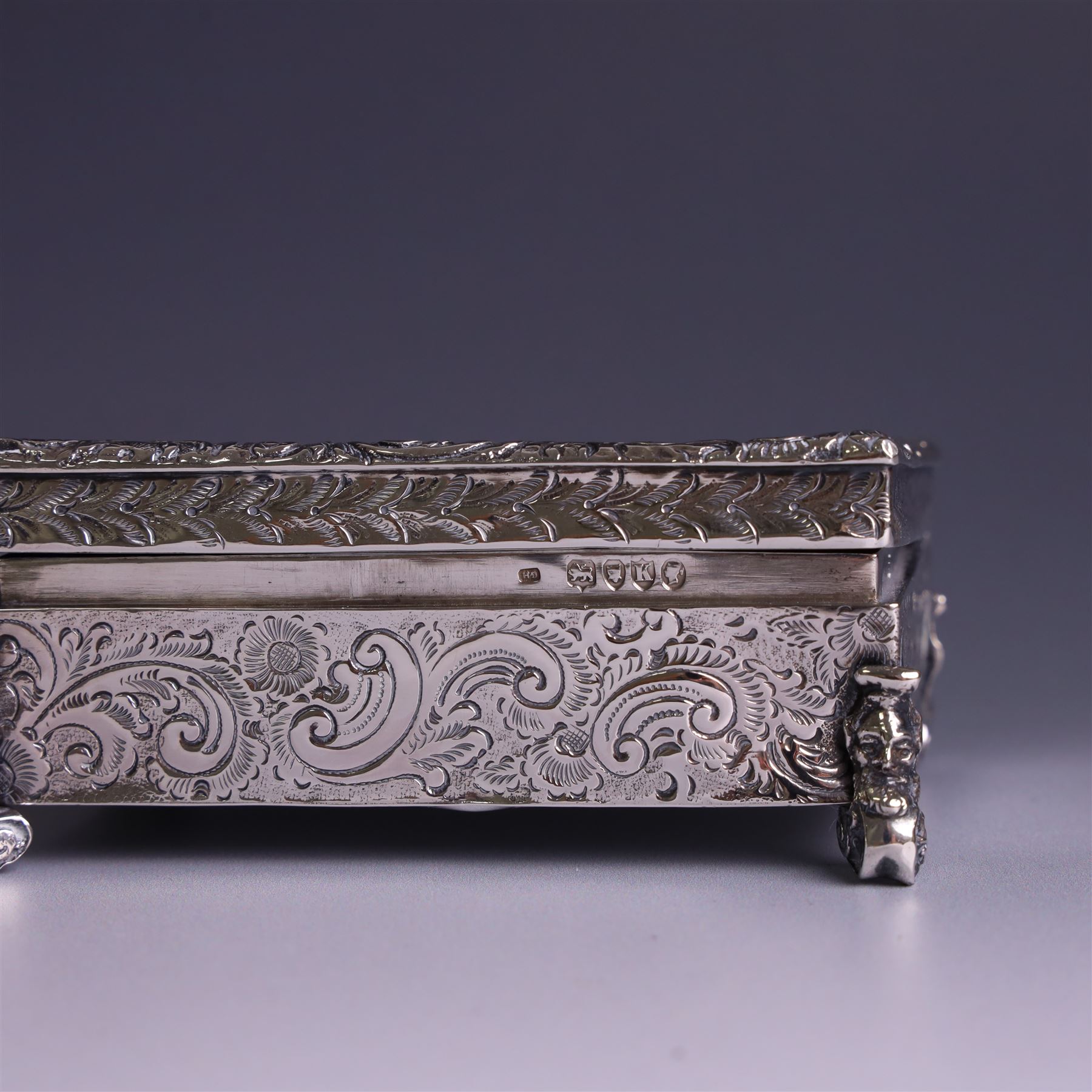 Victorian silver tobacco box, rectangular form, hinged lid embossed with a gaming scene, the body engraved with scrolls and flowers, upon four scrolling mask feet, hallmarked George Heath London 1885, H4.5cm, W10.5cm