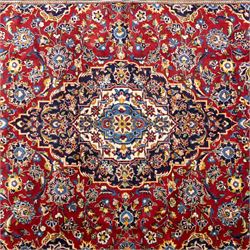 Central Persian Kashan hand-knotted rug, rich red field with a central lobbed medallion surrounded by finely drawn scrolling floral sprays, palmettes, and rosettes, enclosed by a broad dark blue floral border with multiple guard stripes and cotton fringe ends 