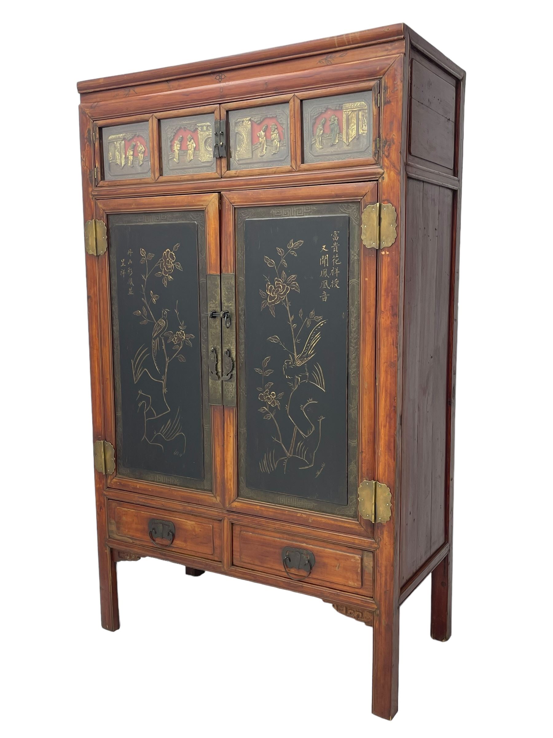 Late 19th century Chinese Qing dynasty red and black lacquered wood cabinet,  Fujianese province, upper cupboard enclosed by two doors with relief carved and gilt panels depicting figures behind glass, the large cupboard enclosed by two panelled doors carved with trailing foliate branches and birds, inscribed with Chinese characters, to the left ‘The red mountains are in the shape of Phoenixes and appear auspicious’ and to the right, ‘Wealth and honour are given by flowers and I hear the sound of the phoenix', fitted with two drawers, on square supports with carved brackets 