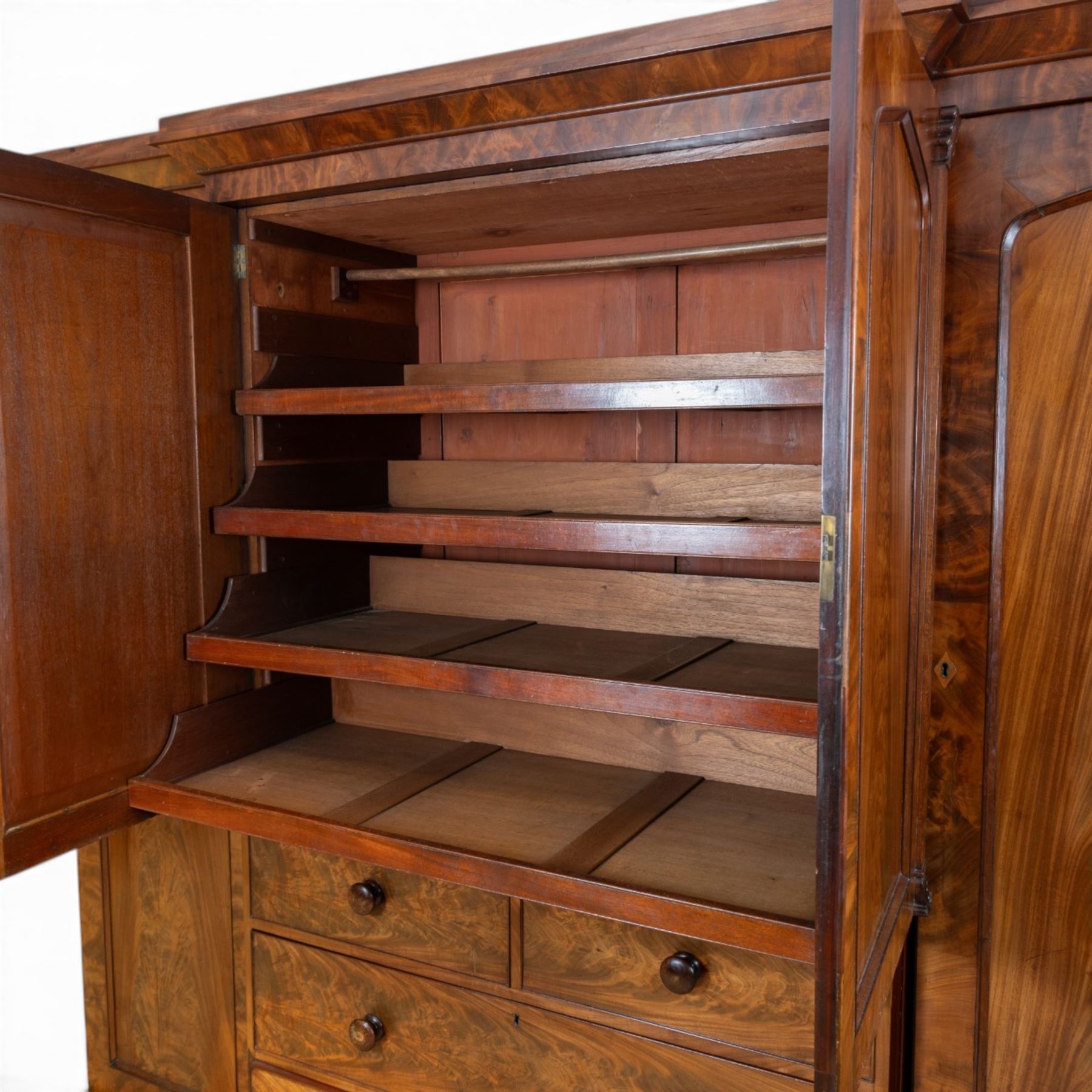 Early 19th century figured mahogany combination wardrobe, the breakfront architectural cornice over central press cupboard enclosing linen slides, flanked by full-height hanging cupboards with arched inset panels, lower arrangement of two short and two long drawers with turned knob handles, applied acanthus carved detail, raised on bun feet