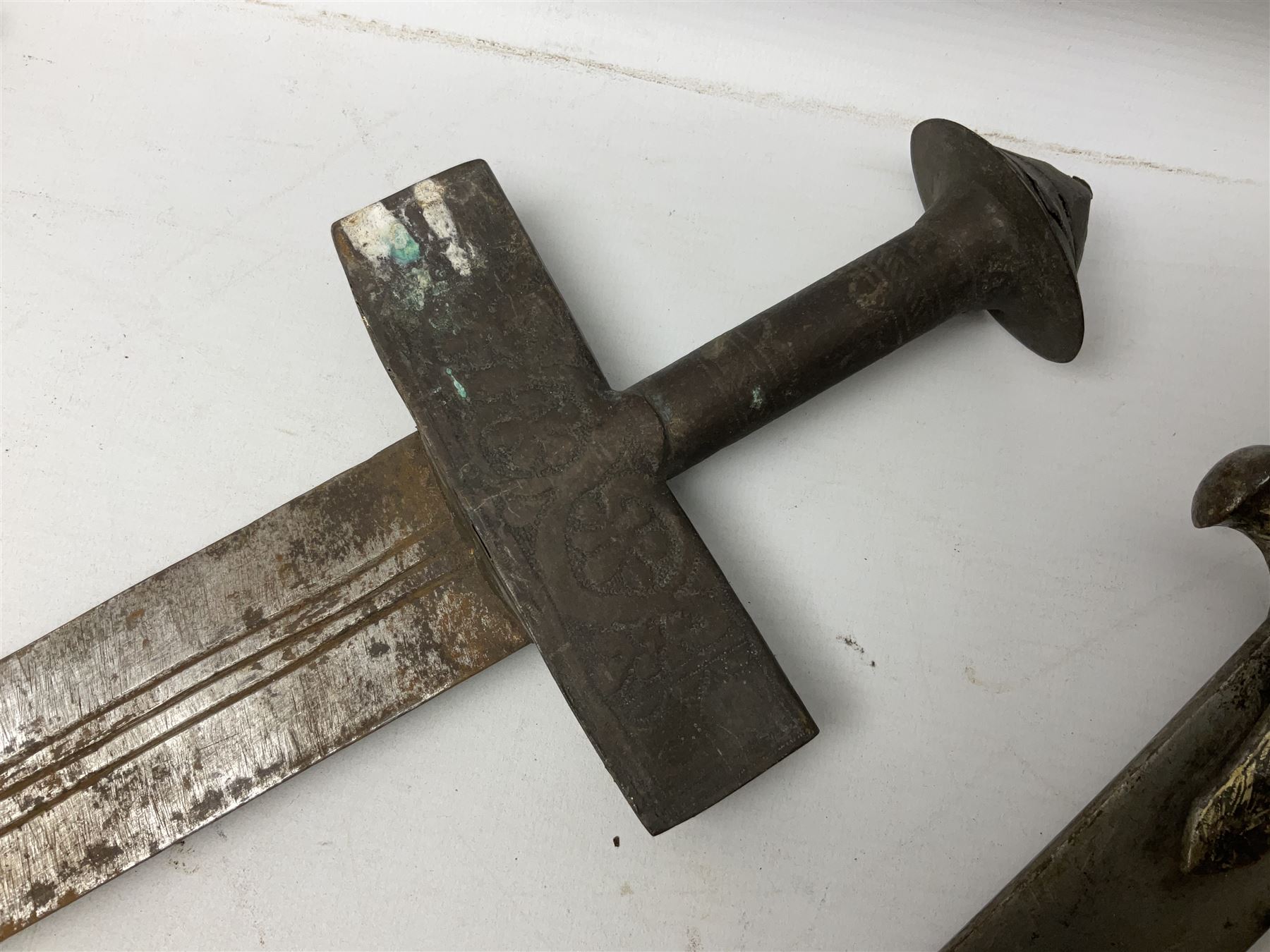 Tuareg Takoba sword with 78.5cm triple fullered blade incised with two crescent moons to either side, metal hilt with stylised floral designs to the cross-guard, decorative grip and disc form pommel cap; in tooled leather scabbard decorated with geometric designs L92cm overall; and an Indian Tulwar sword with 75cm plain curving blade and 'silver' damascened hilt (no scabbard) (2)