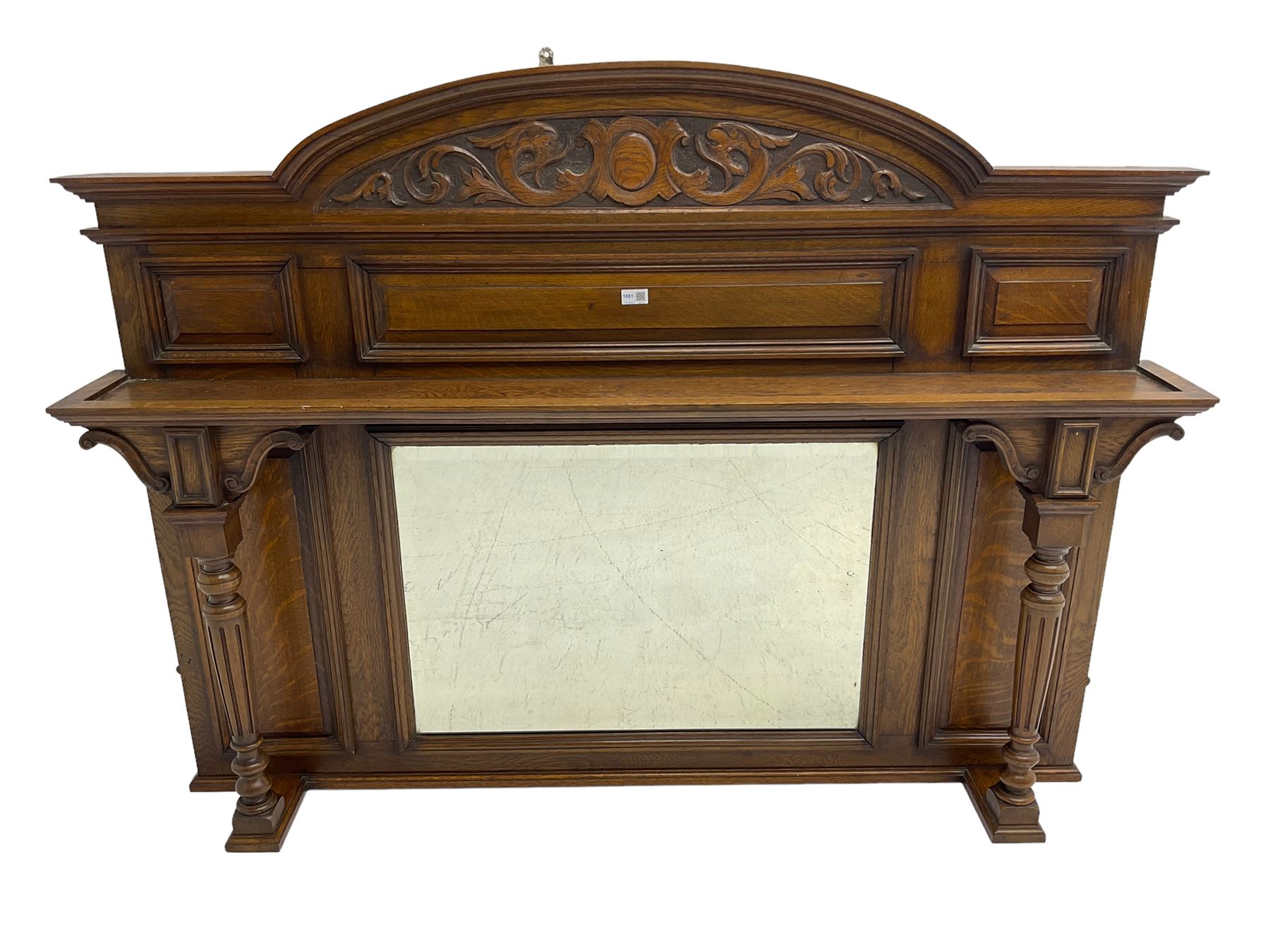 Oak overmantle mirror, arched pediment carved with shield and flanking fish with trailing scrollwork, panelled frieze over extending canopy raised by turned and fluted column pilasters, bevelled mirror plate 