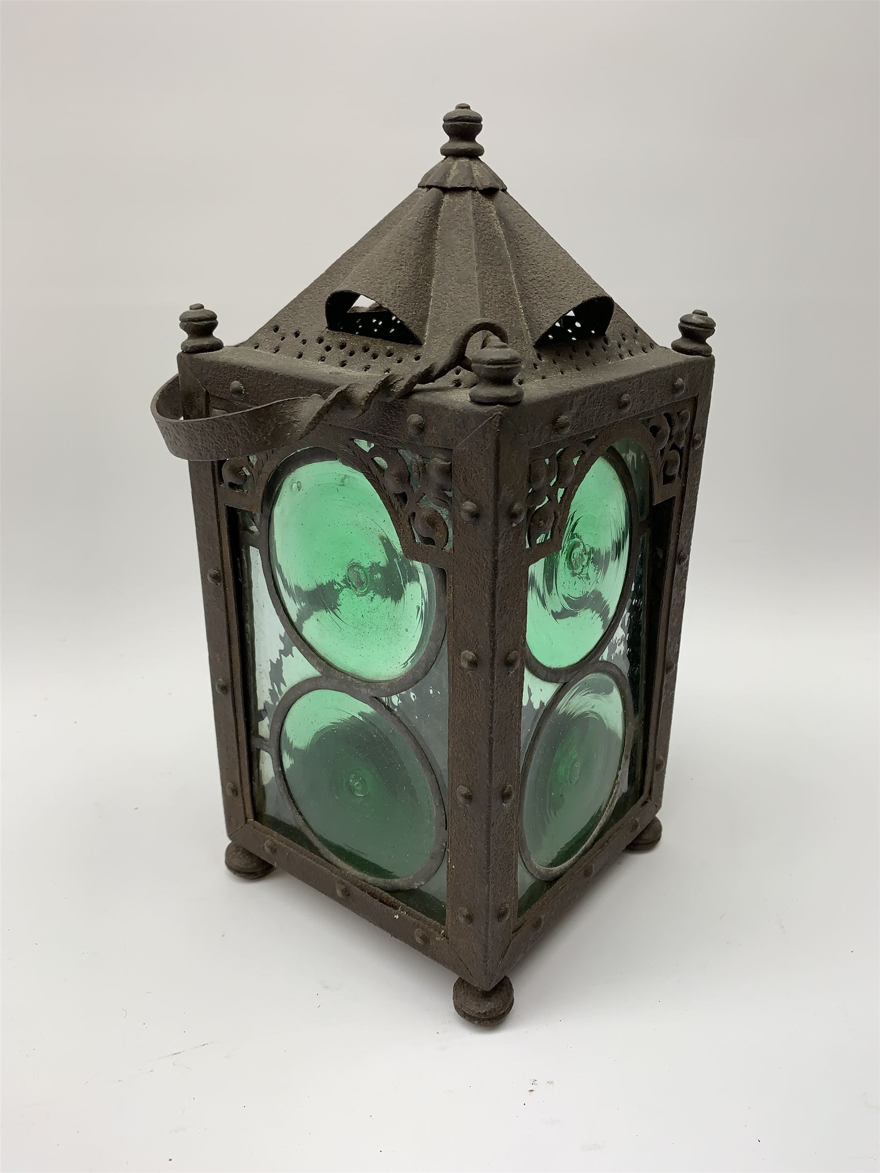 An Arts & Crafts wrought iron lantern, of square sided form with swing handle, and green and clear glass panels, raised upon four compressed bun feet, not including handle H30.5cm.