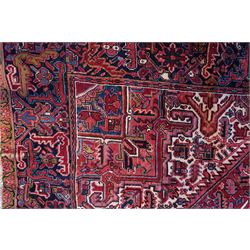North West Persian Heriz hand-knotted carpet, brick-red field with a large central angular star medallion with pendant motifs, surrounded by stylized geometric palmettes, serrated leaves and tribal devices, ivory corner spandrels similarly decorated, field enclosed by a dark indigo main border of stylized rosettes and angular vine motifs between multiple guard stripes, with cotton fringe ends  