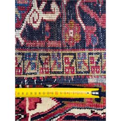 Northwestern Persian Heriz hand-knotted carpet, red madder field with a bold stepped medallion in indigo, ivory, and terracotta, flanked by angular floral motifs and stylized leaves, the spandrels set within a dark indigo border of rosettes and angular vine motifs between narrow guard stripes, 340cm x 245cm 