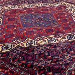 North West Persian Senneh hand-knotted rug, dark indigo field with a central bold stepped lozenge medallion of madder red and deep blue, enclosed within an ivory serrated contour forming a cruciform outline, medallion with repeating boteh and floral devices, field decorated with an all-over Herati pattern of rosettes enclosed by angular leaves principal border of madder red ground decorated with alternating floral heads, flanked by narrow guard stripes, with cotton fringe ends 