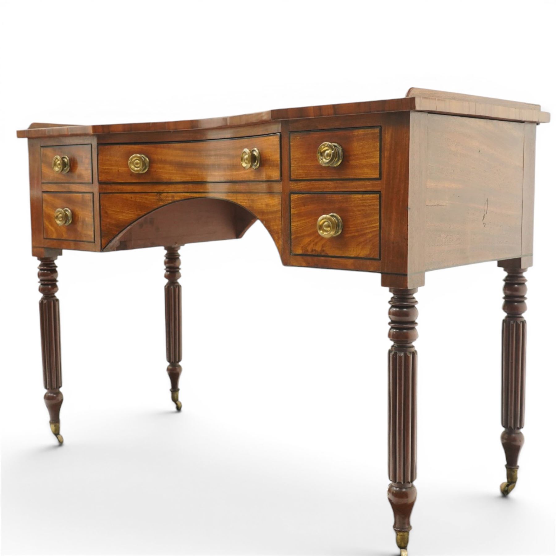 In the manner of Gillows - Regency period mahogany kneehole dressing table or writing desk, the shaped rectangular top with raised three-quarter gallery, fitted with central concave frieze drawer flaked by two graduating drawers to each side, each with ebony stringing, raised on turned and reeded supports terminating to brass cups and castors