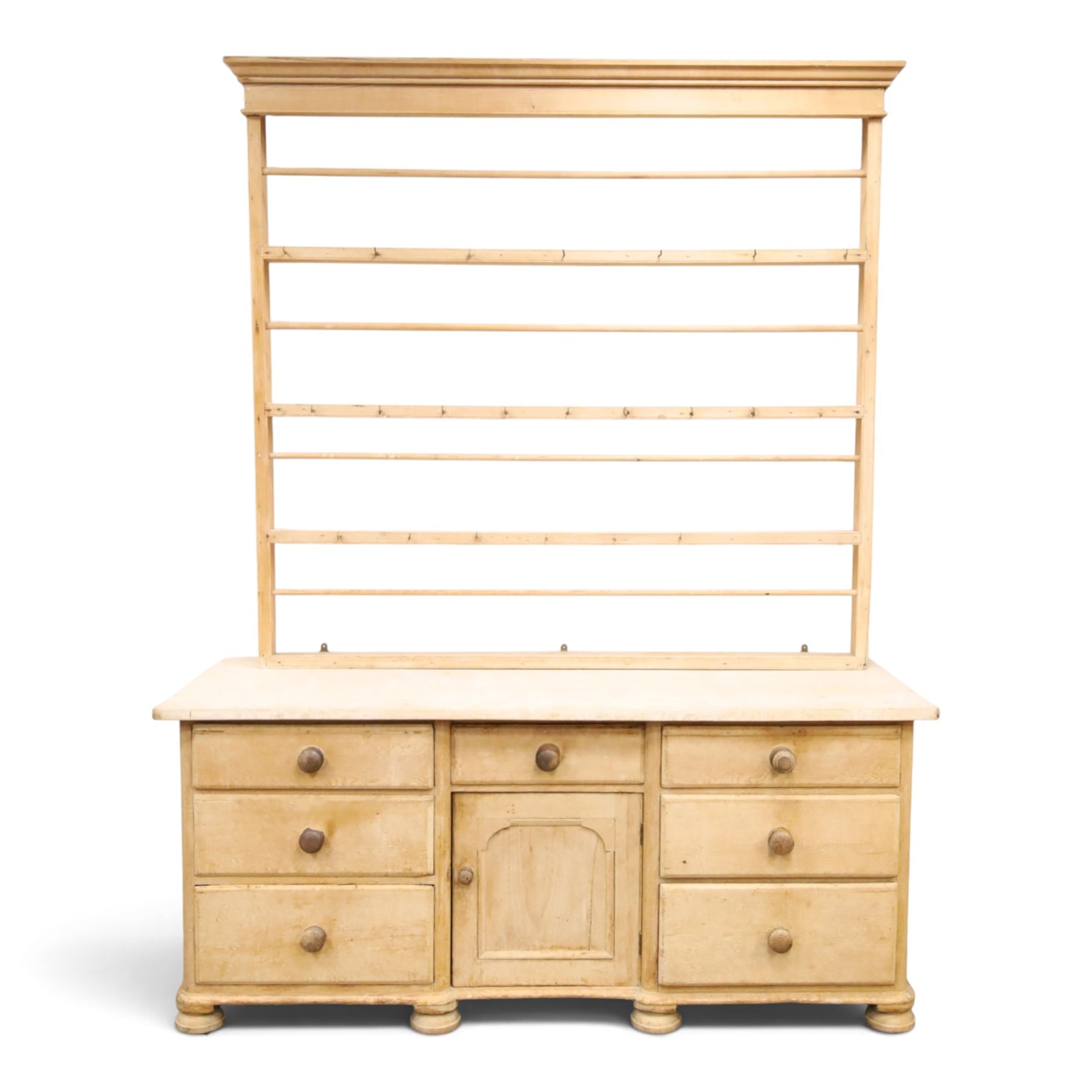 Victorian stripped pine dresser base with plate rack, the four-tier plate rack with supporting rails and hooks, the inverted-breakfront base fitted with seven assorted graduating drawers surrounding a recessed cupboard, on compressed bun feet with castors