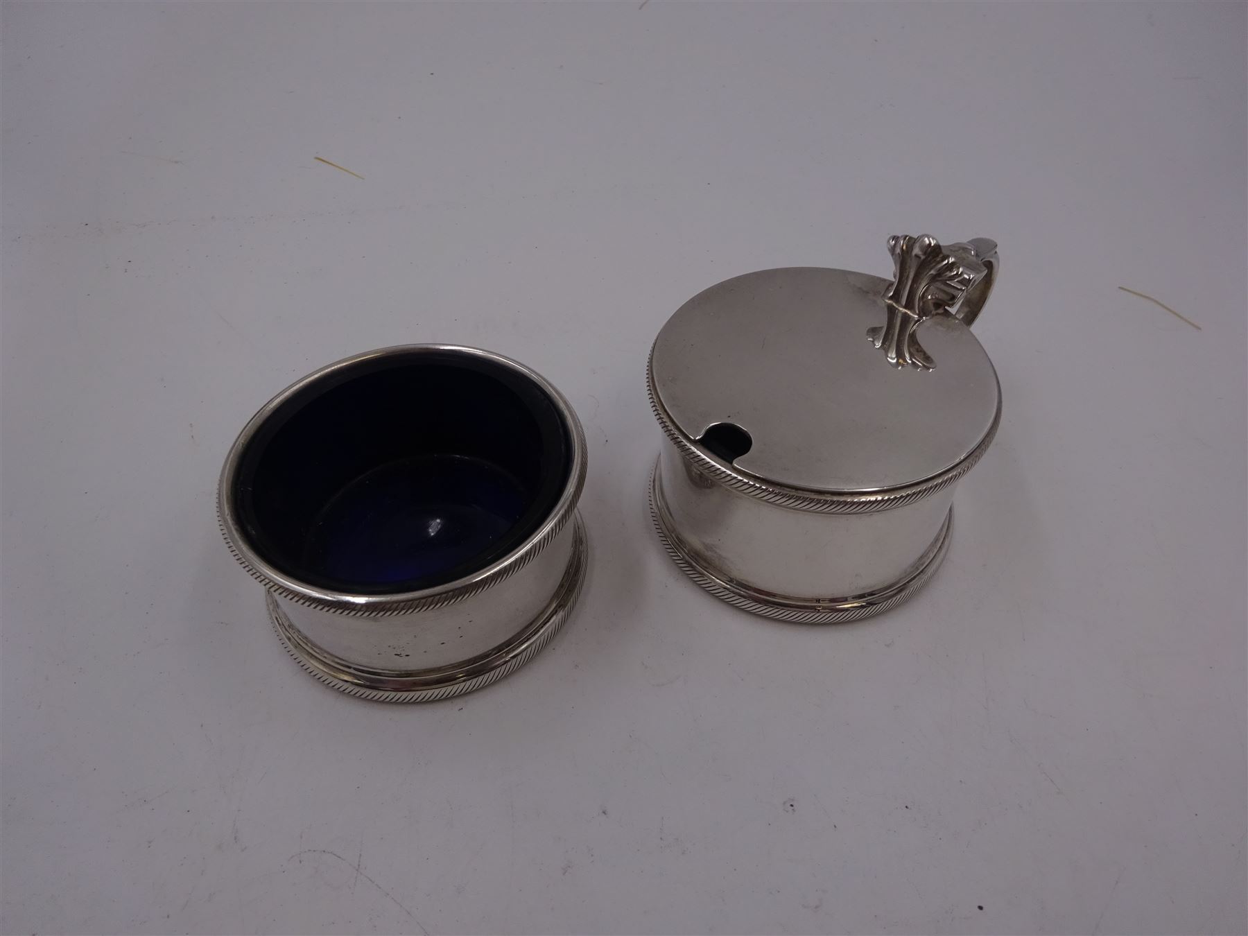 1930s silver part cruet set, comprising mustard pot and cover and open salt, both of circular drum form with oblique gadrooned rim, the mustard pot with capped scroll handle and palmette thumbpiece, both with blue glass liners, hallmarked London 1935, maker's mark worn and indistinct
