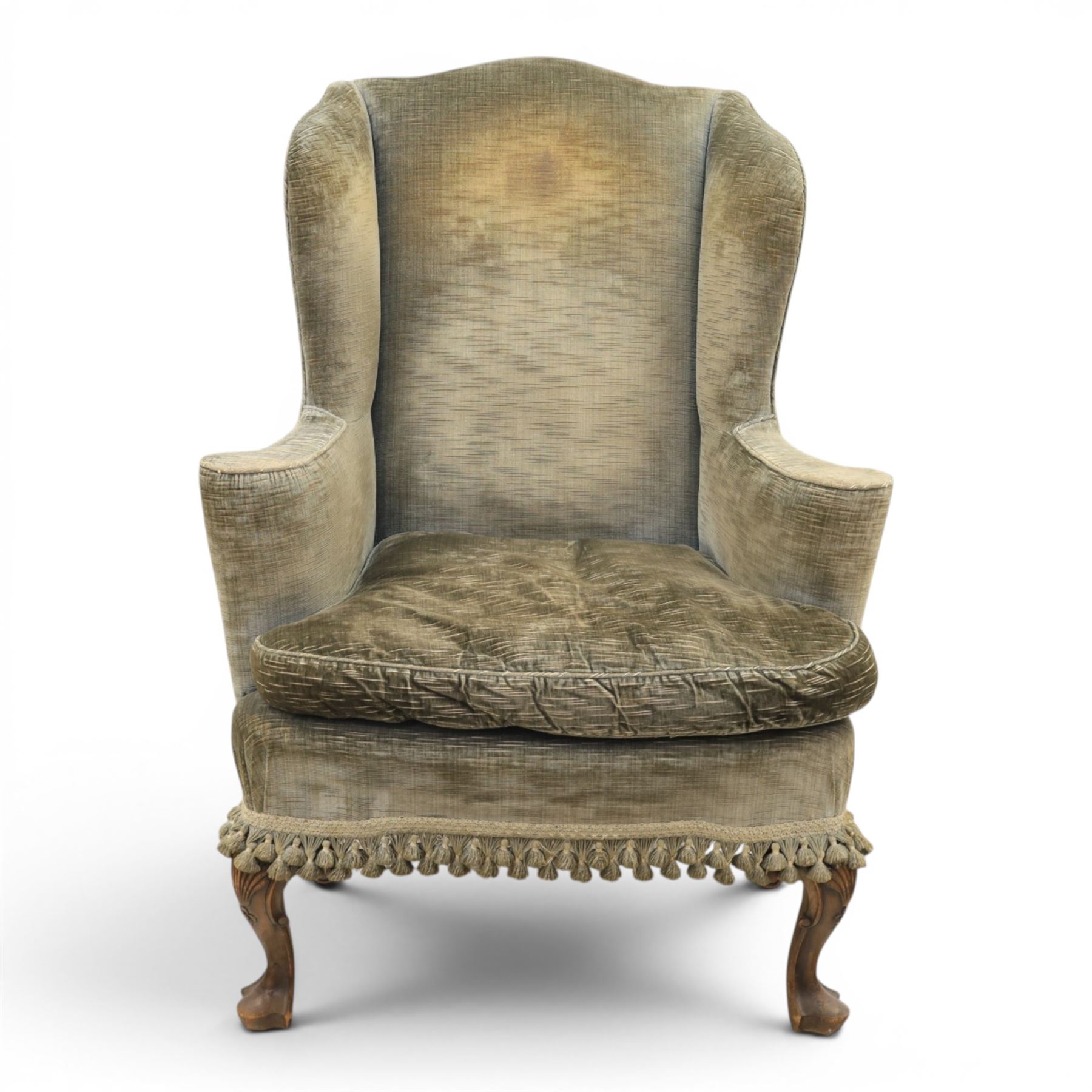 Pair of Georgian style wing armchairs, upholstered in green velvet, each with shaped wing backs, padded scroll arms and loose seat cushions, raised on cabriole supports terminating in pad feet, one with tasselled fringe