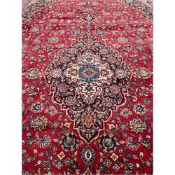 Large Persian Kashan crimson ground carpet, large floral central medallion surrounded by trailing branches decorated with leaves and stylised plant motifs, the main border with scrolling design interspersed with palmettes, within floral pattern guard stripes 